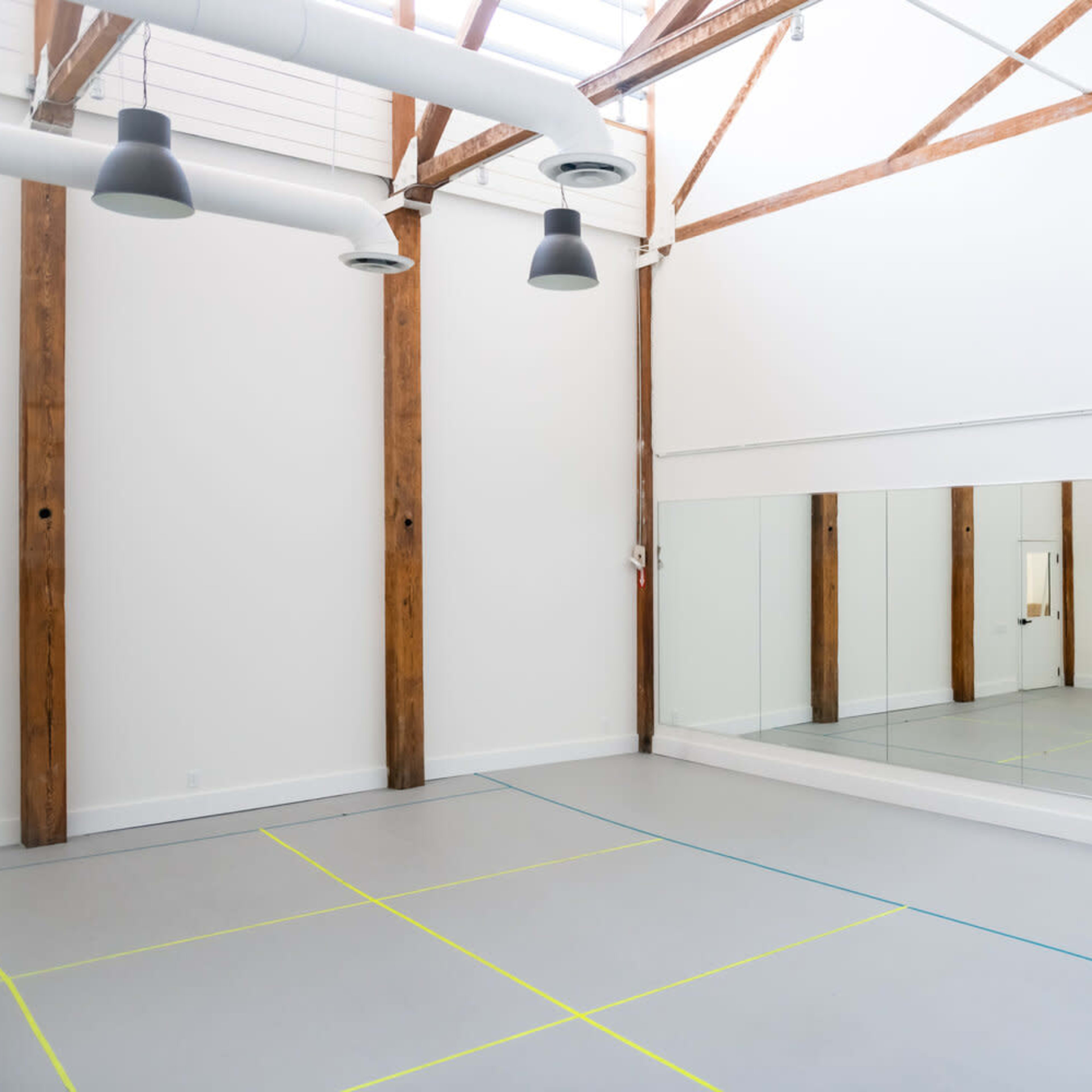 A spacious dance studio features high wooden beams, a mirrored wall, and a light gray floor marked with colored lines.
