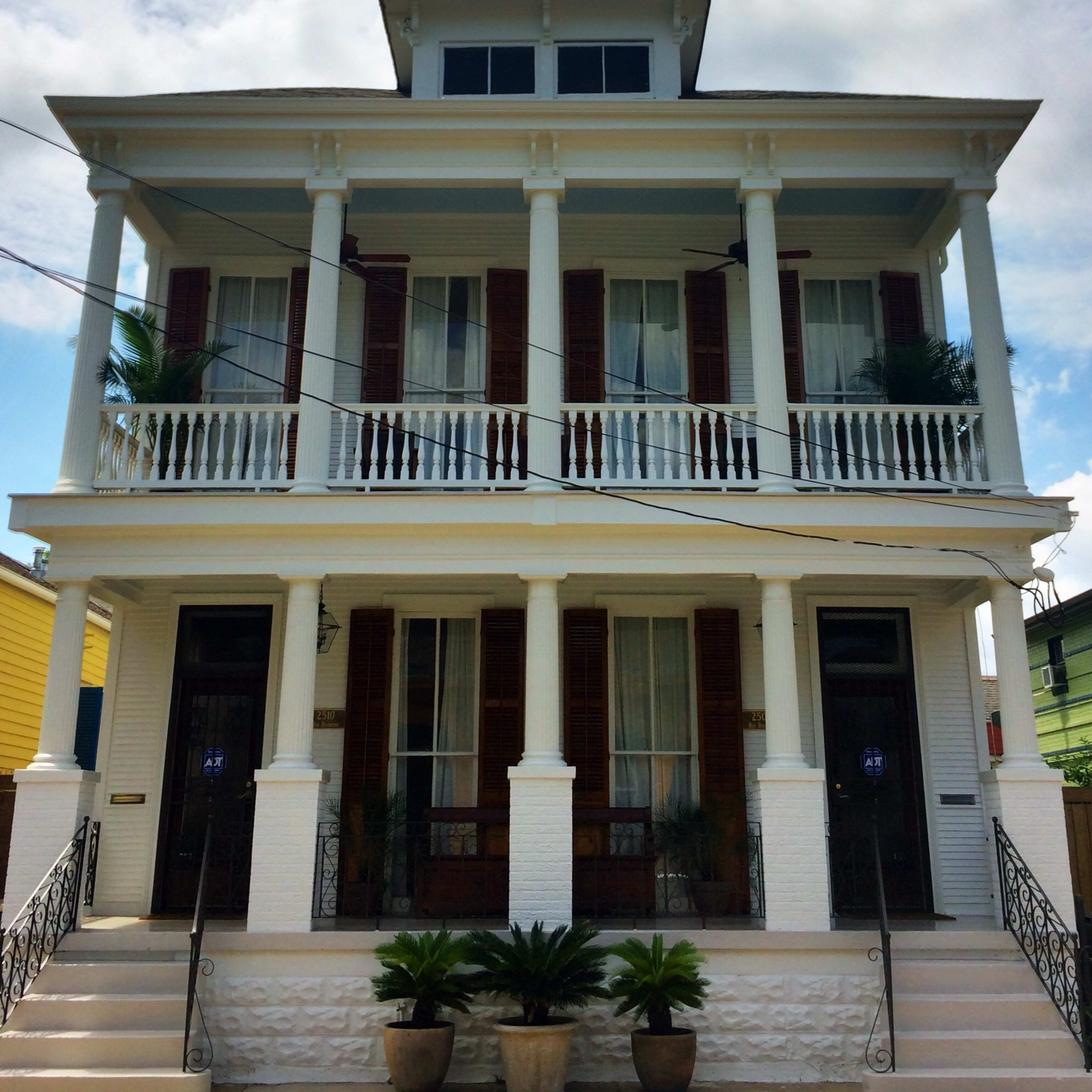 A two-story white house with a porch supported by six columns, featuring wooden shutters and potted plants at the entrance.