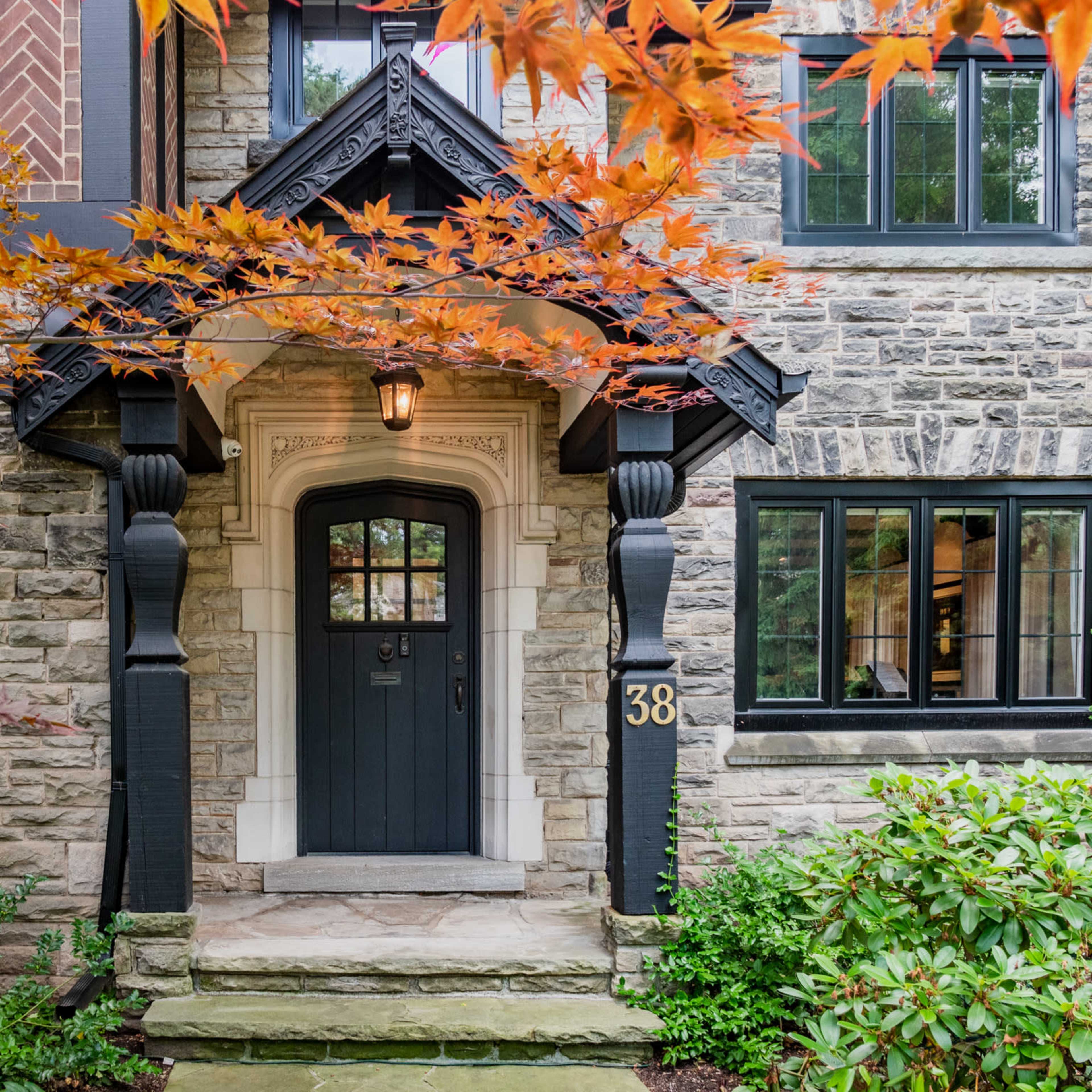 The entrance of a stone-clad house features a dark door framed by ornate woodwork and flanked by lush greenery and red maple leaves.
