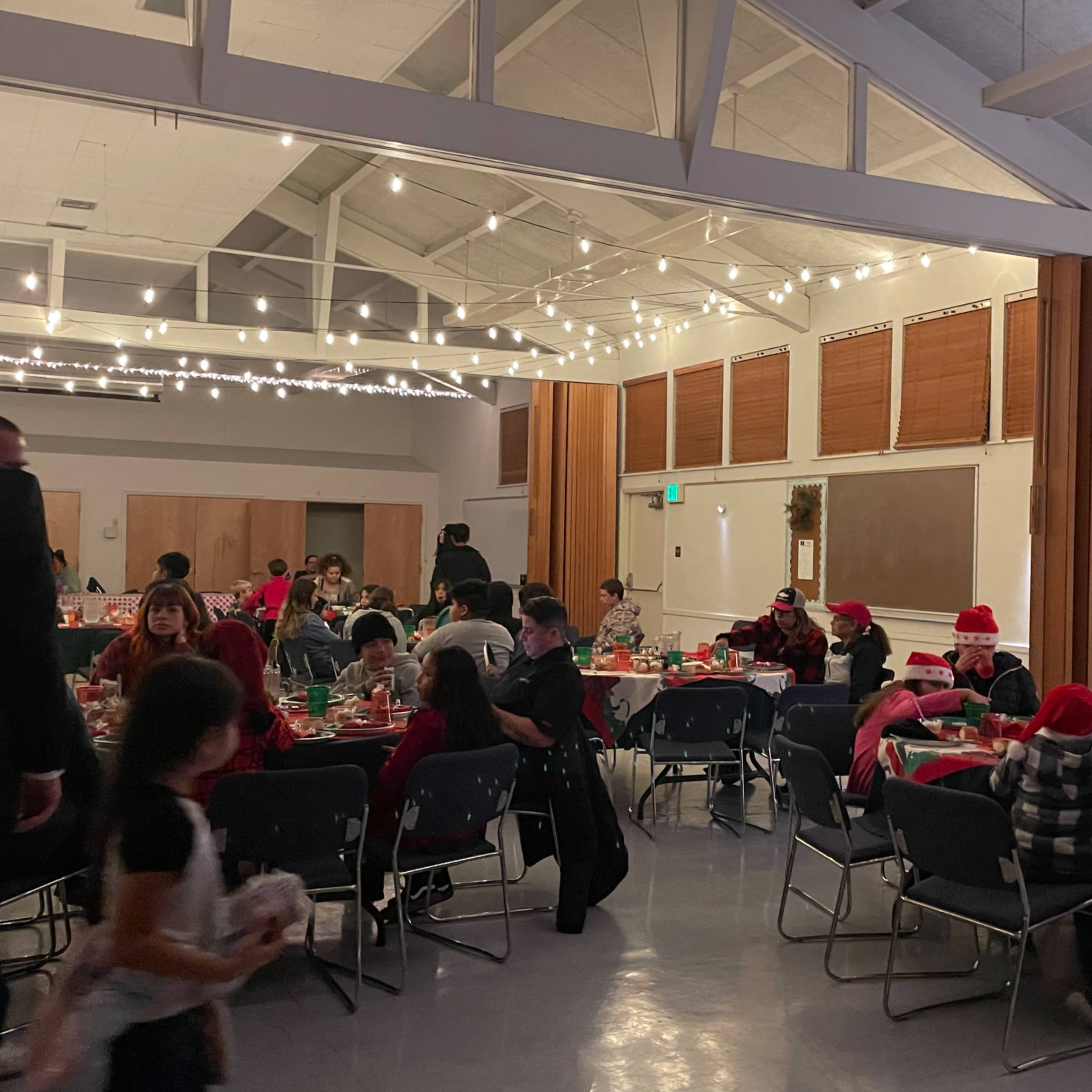 A group of people wearing Christmas hats gathers around tables decorated for a holiday party, with string lights illuminating the room.