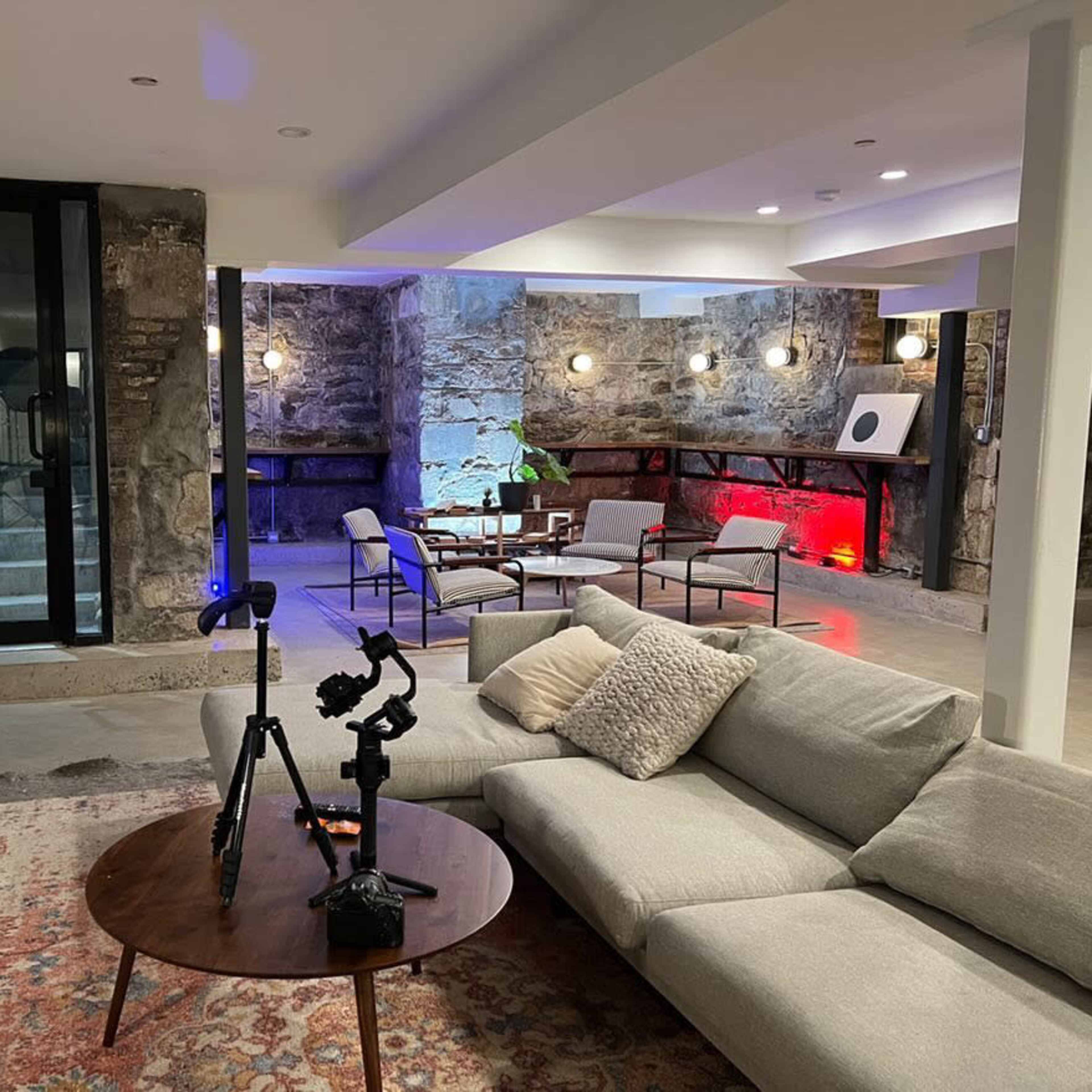 A modern basement living area features a gray sectional sofa, a round wooden coffee table, various seating arrangements, and exposed stone walls illuminated by colored lights.