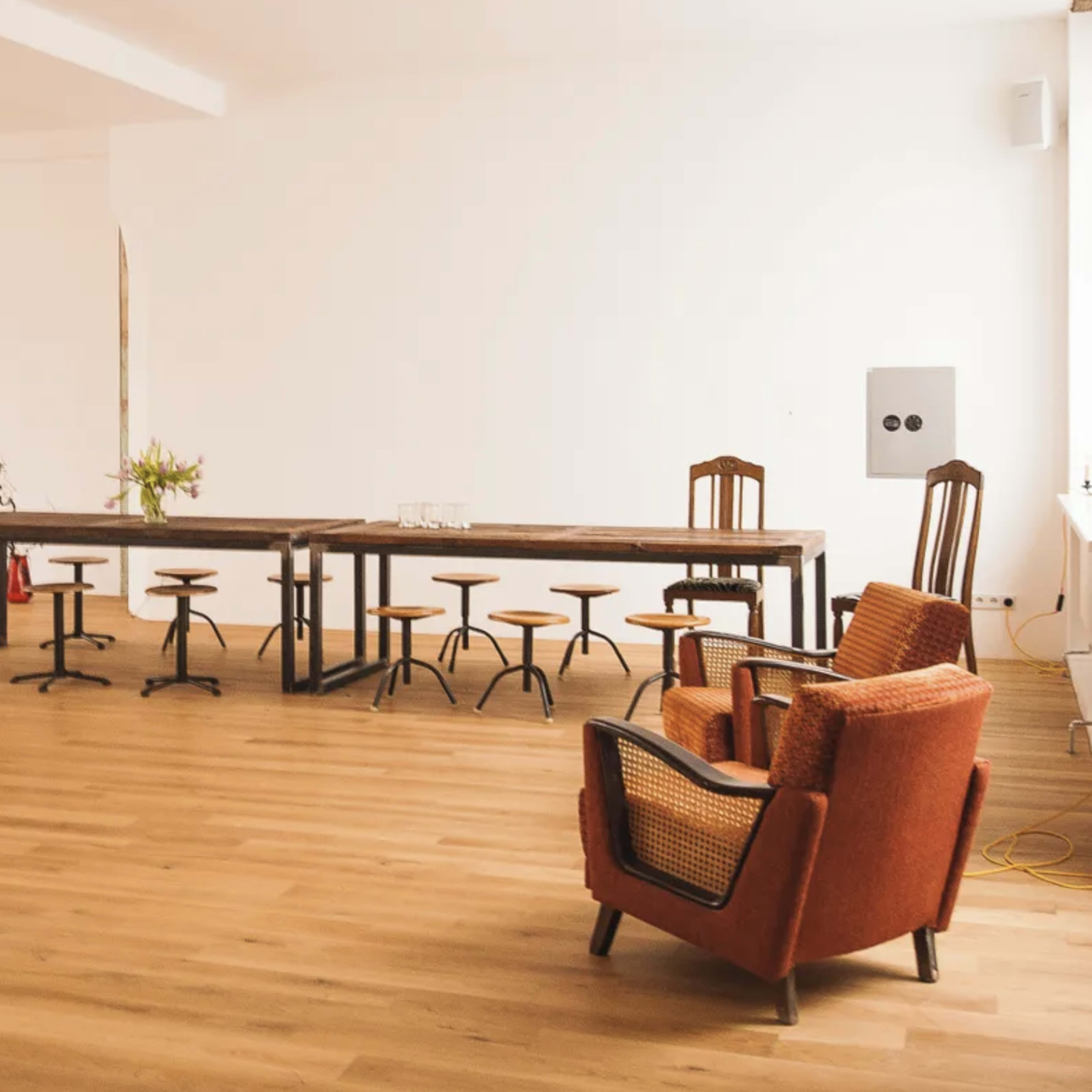 A minimalist interior space featuring a large wooden table surrounded by several stools and an orange armchair at the side.