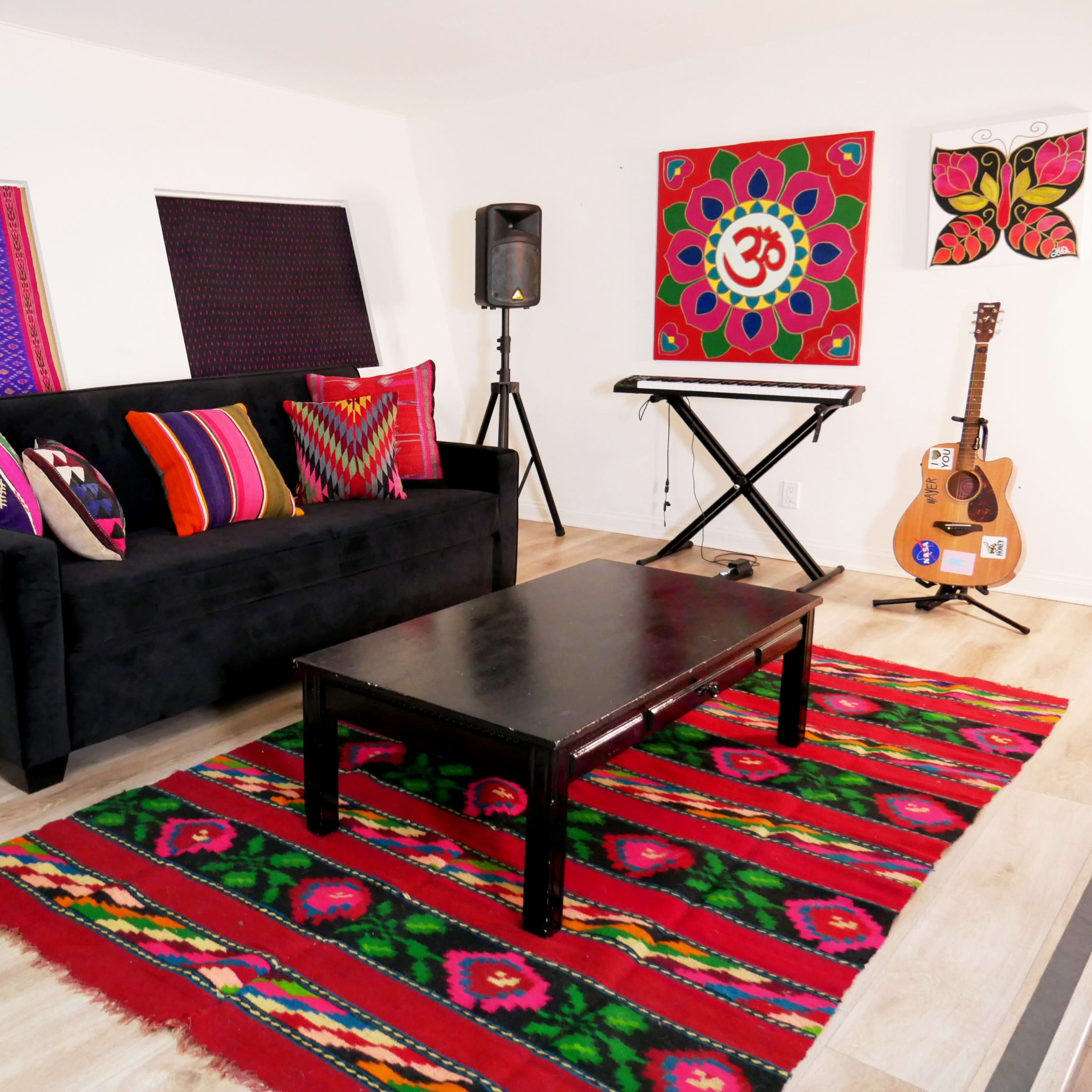 A colorful living room with a black sofa adorned with patterned cushions, a wooden coffee table, a red patterned rug, a guitar, a keyboard, and decorative wall art.