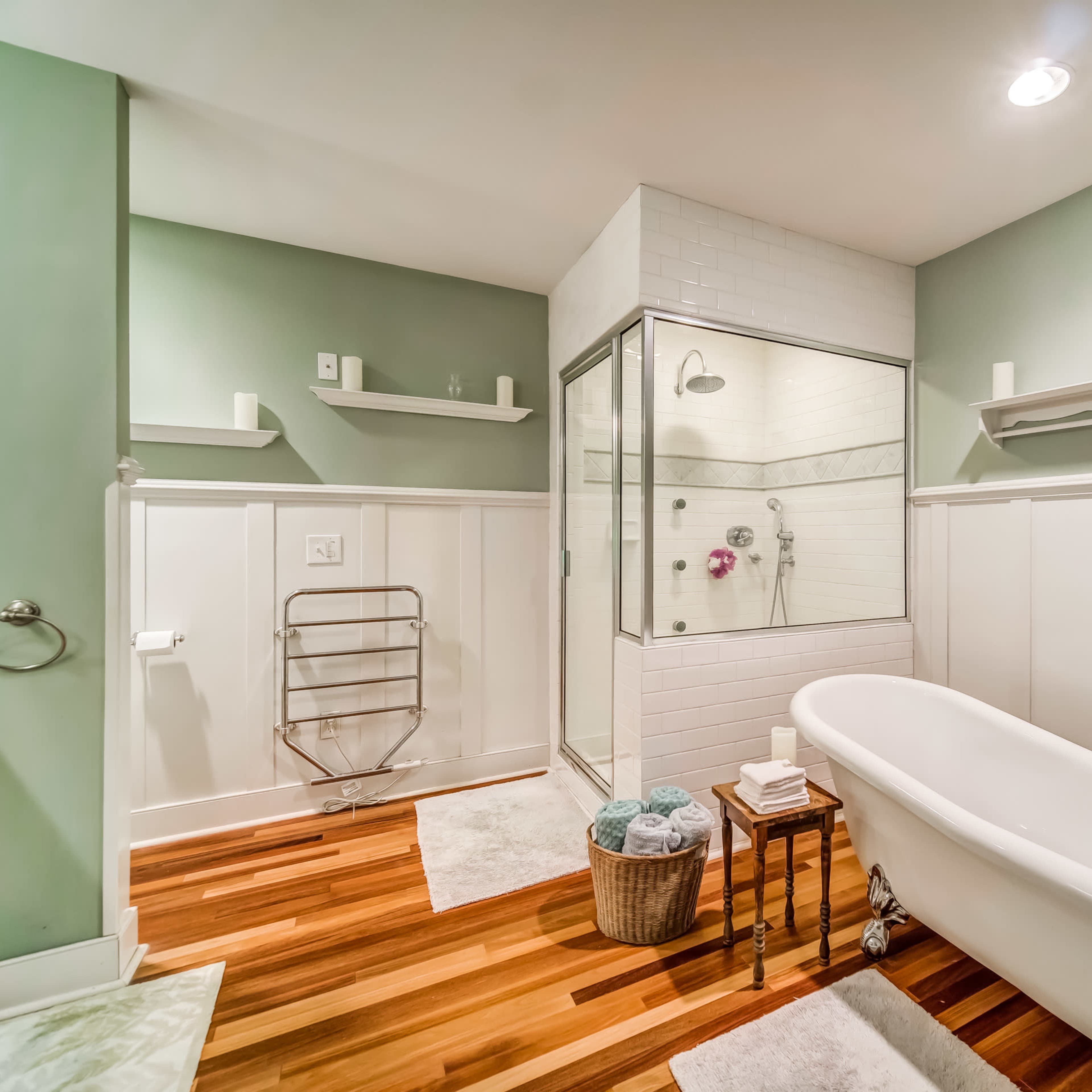A modern bathroom featuring a freestanding bathtub, a glass shower enclosure, wooden flooring, and light green walls with white paneling.