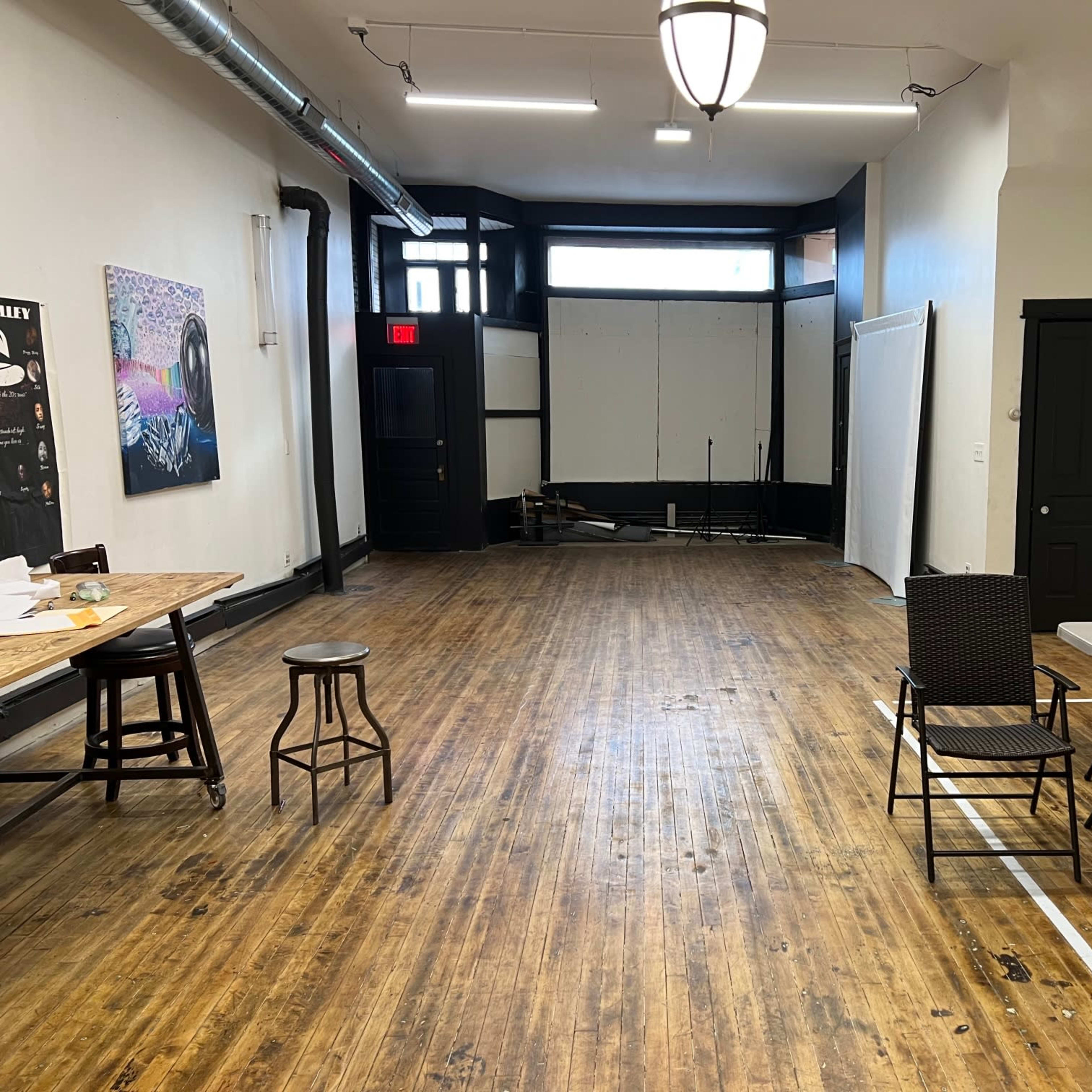 A spacious, empty room with wooden floors, a few tables and chairs, and large windows providing natural light.