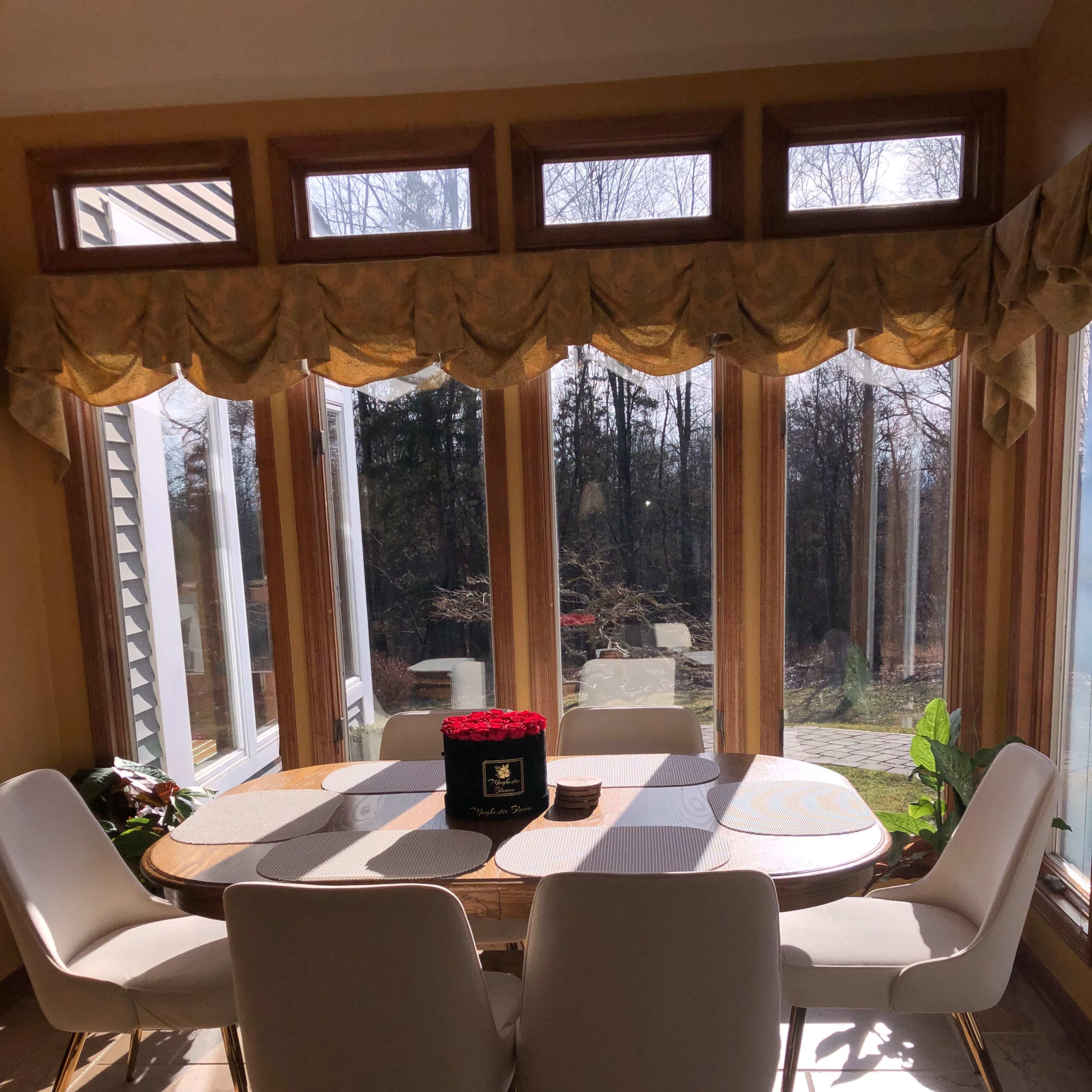 A dining table with six white chairs is positioned in a sunlit area with large windows, revealing a view of a garden.
