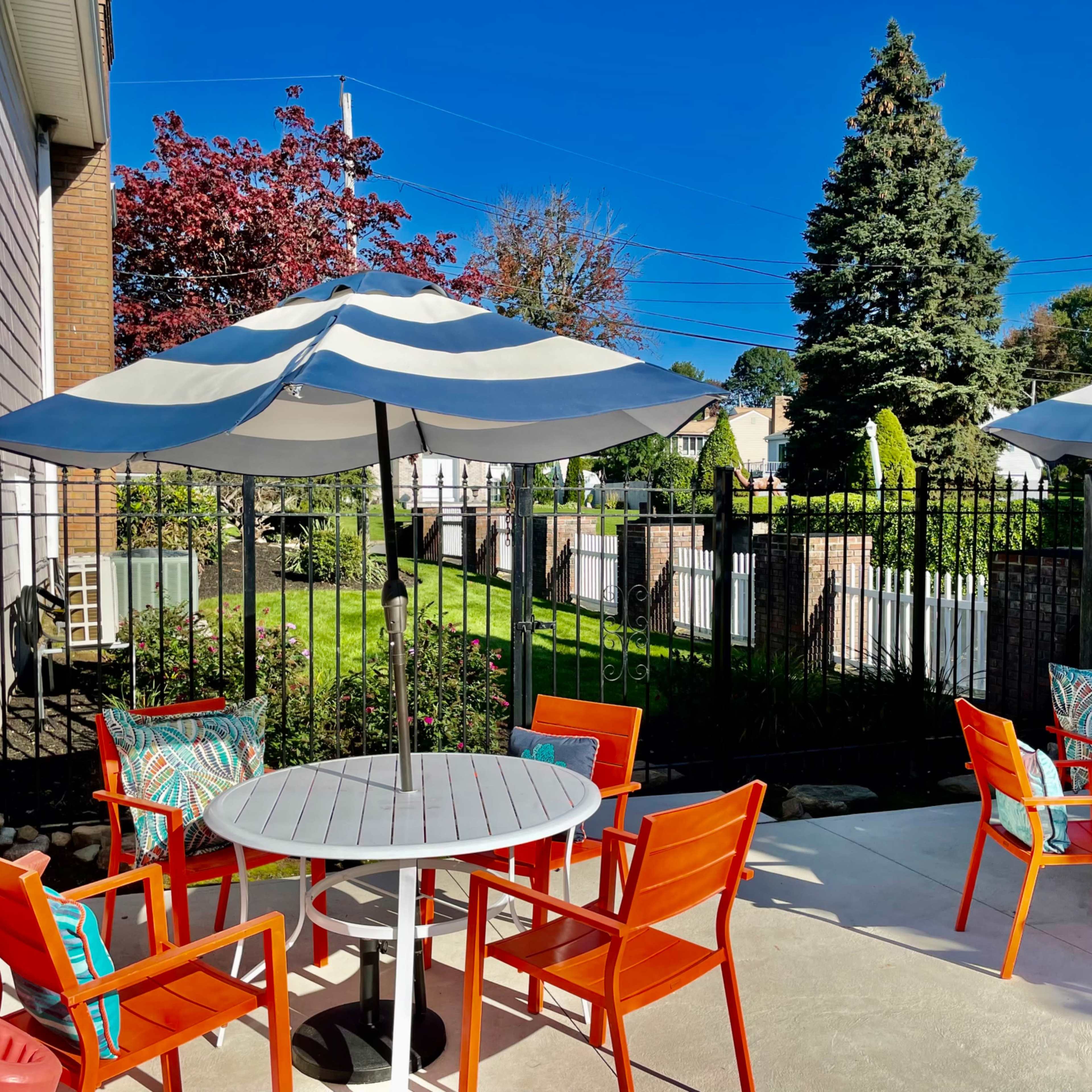 A patio area features round tables with striped umbrellas and orange chairs, surrounded by green grass and trees.