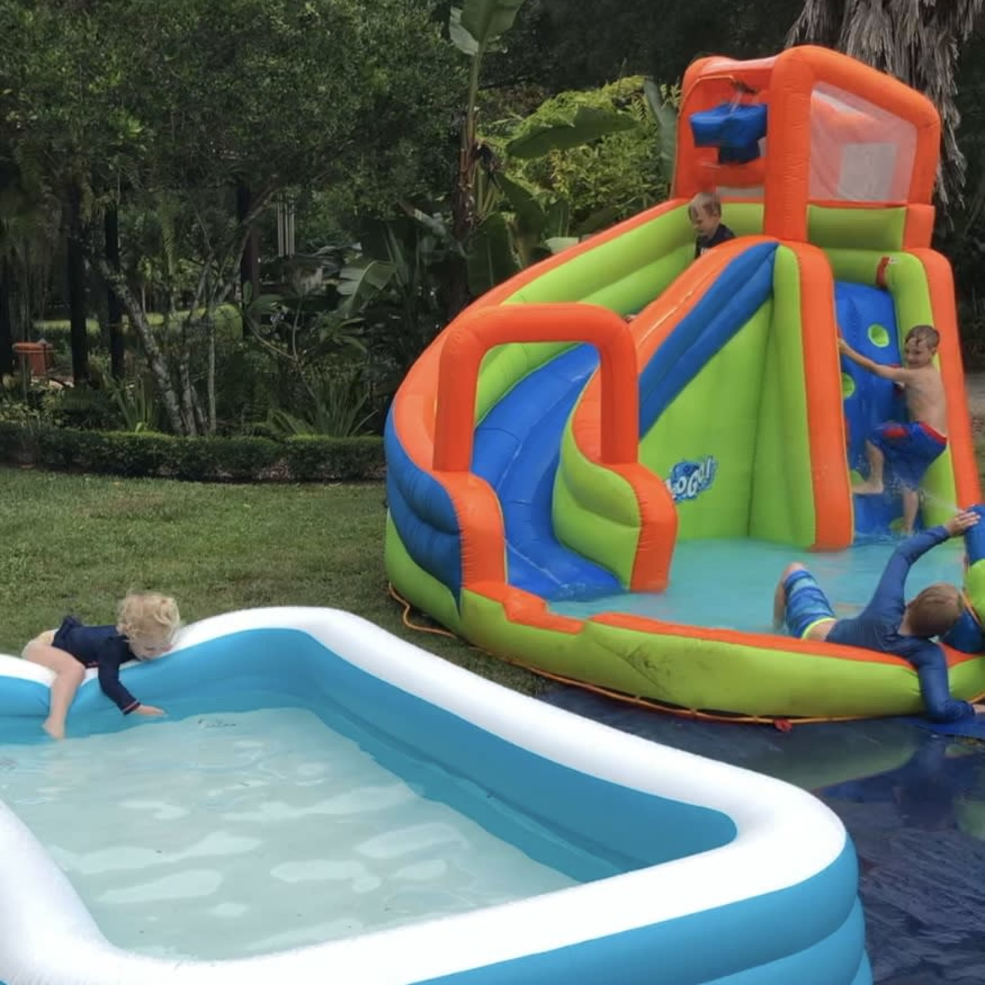 Children are playing on a colorful inflatable water slide and in a small inflatable pool in a lush, green backyard.