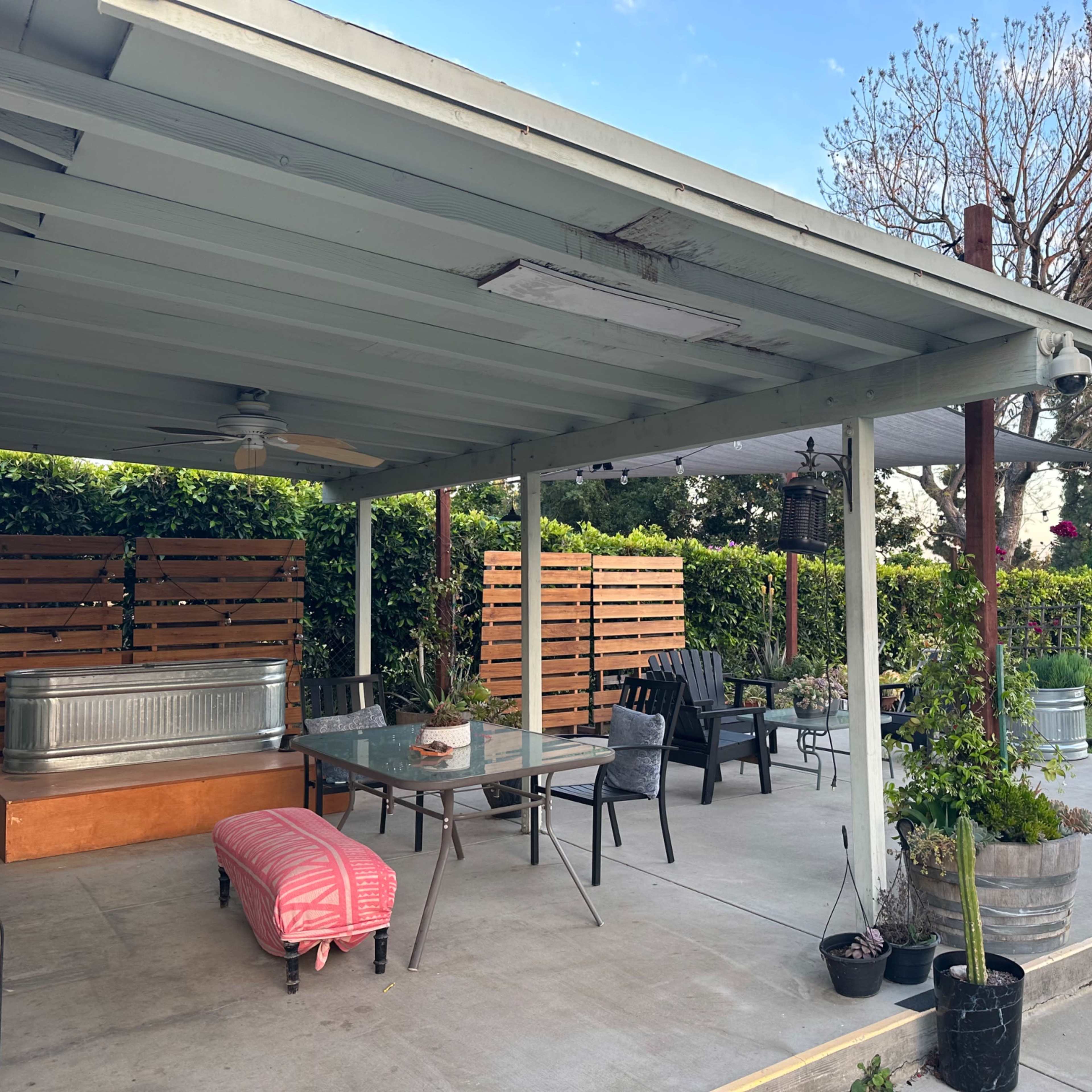 A covered outdoor patio area with a table and chairs, surrounded by plants and wooden privacy panels.