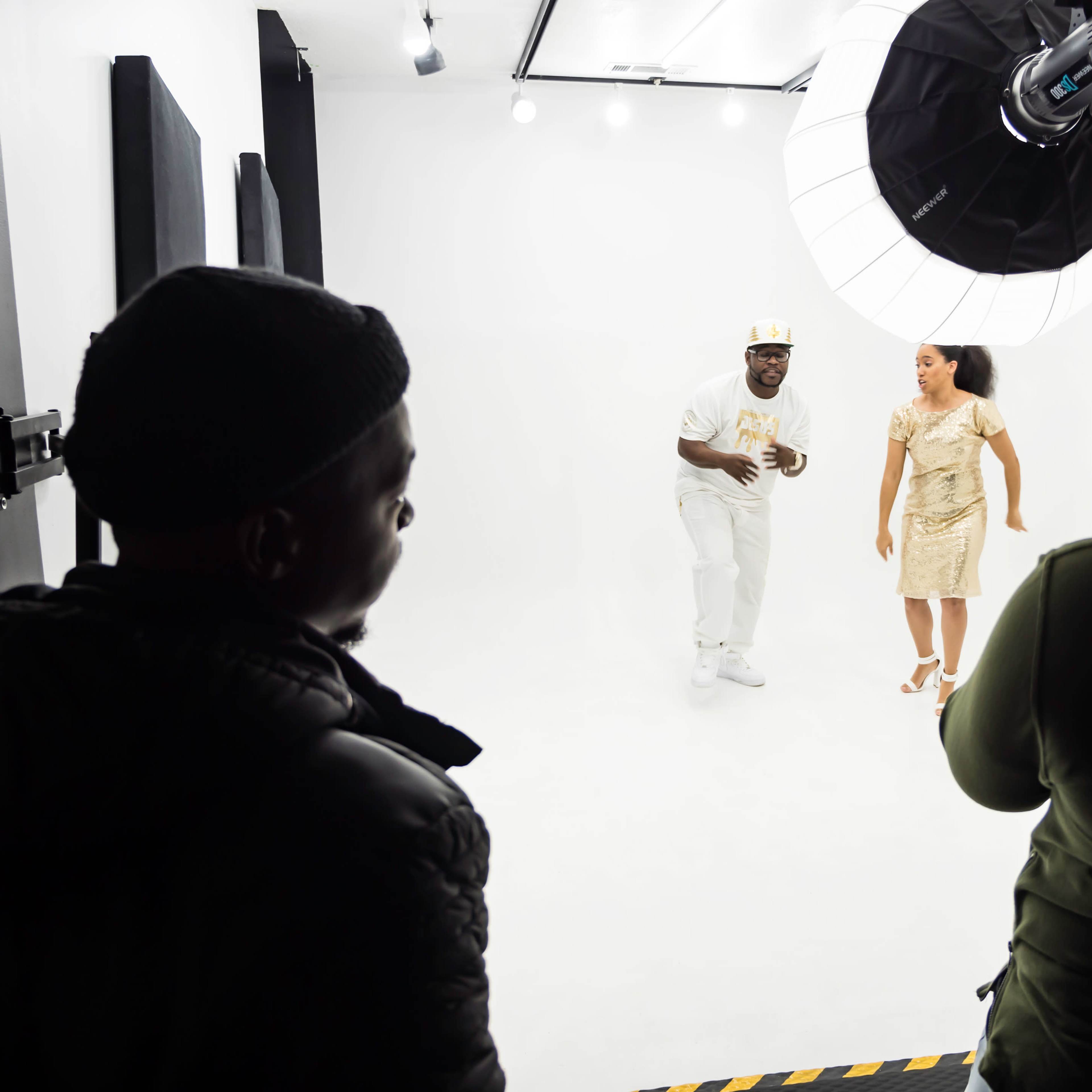 A man and a woman are posing in a photography studio with white backdrop lights while two people are observing and capturing the moment.