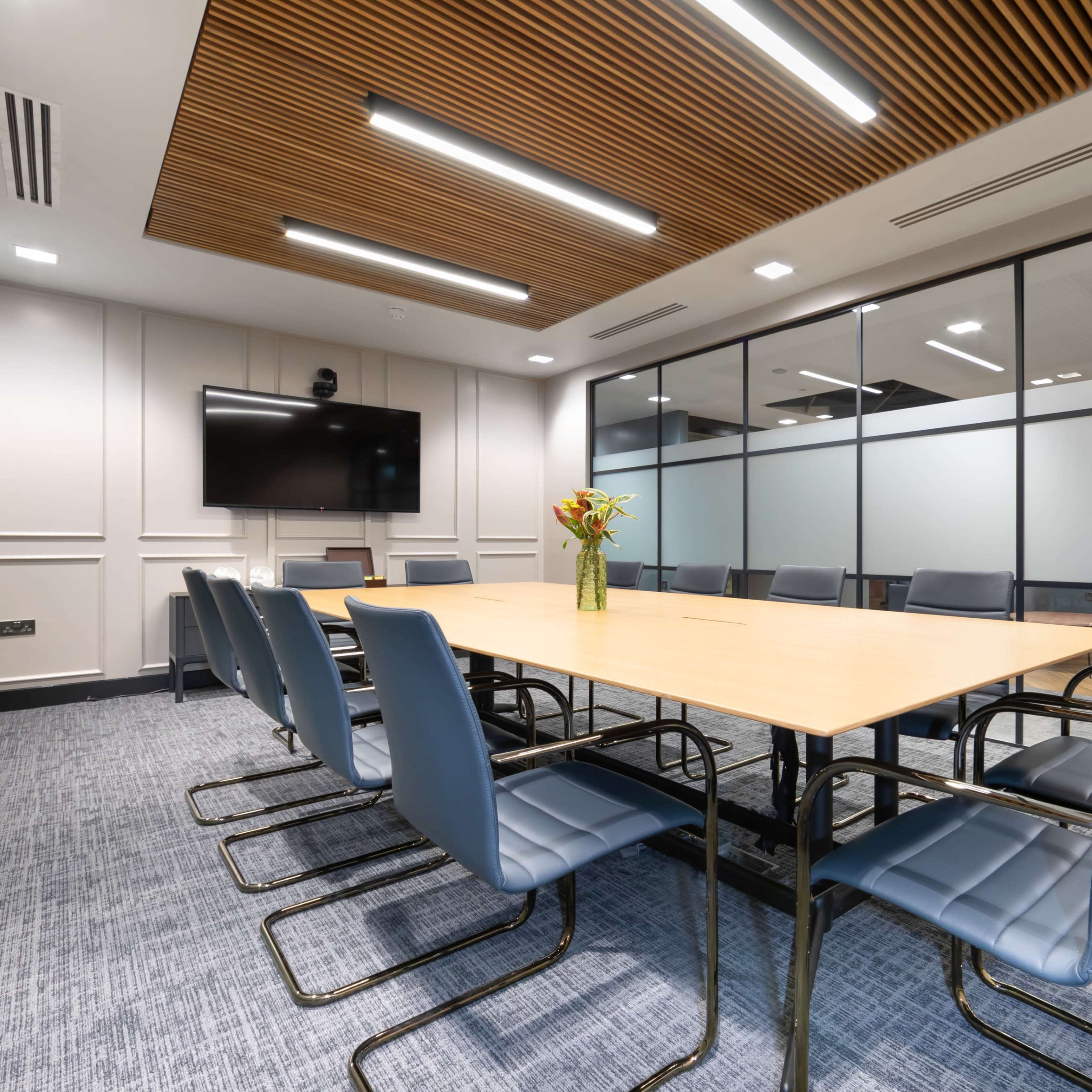 A modern conference room features a large rectangular table surrounded by metal-framed chairs, with glass walls and a television mounted on one side.