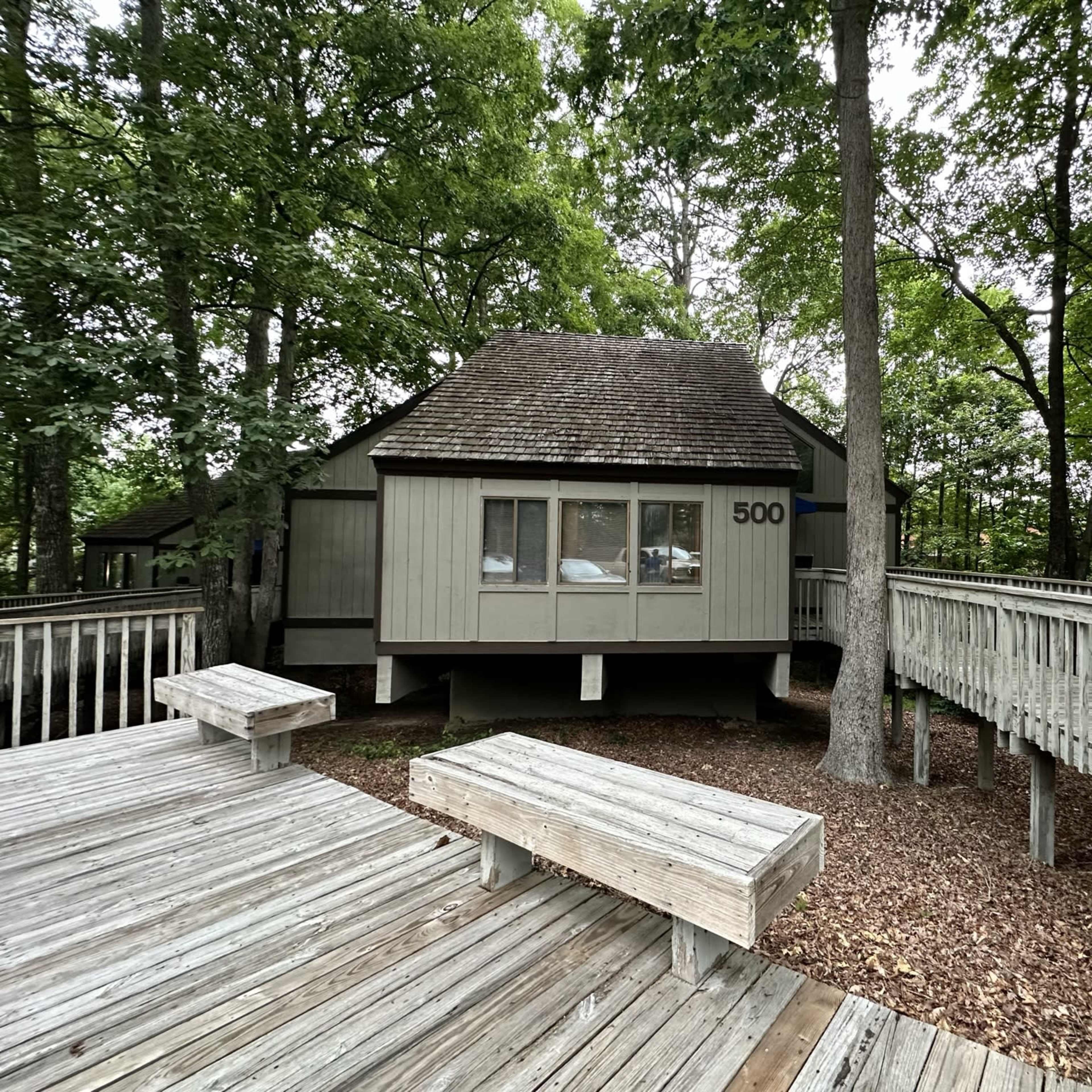 A cabin with the number 500 is situated among trees, connected by wooden walkways and surrounded by a deck area covered in wood chips.