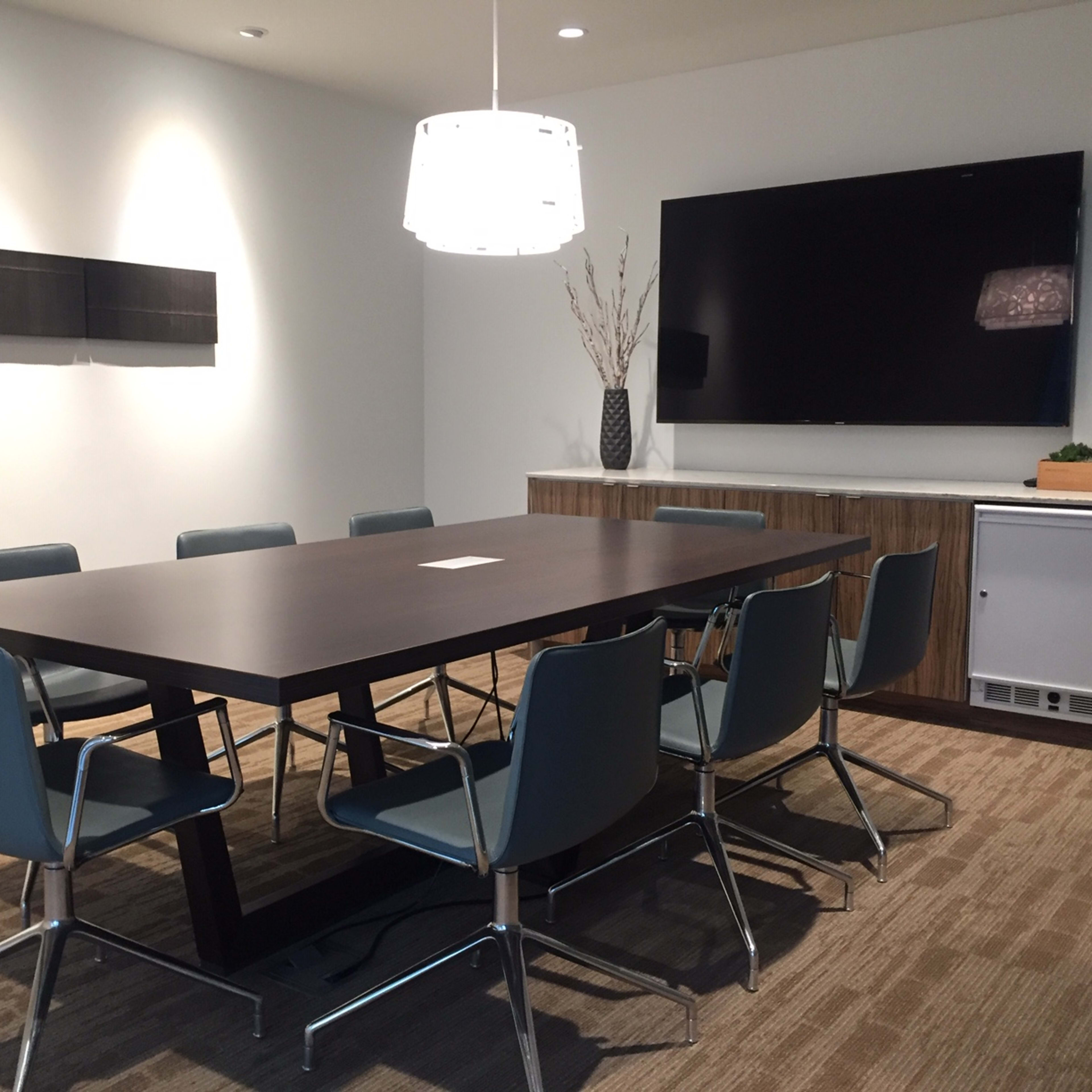 A conference room features a large rectangular table surrounded by six blue chairs, a wall-mounted television, and minimal decorative elements.