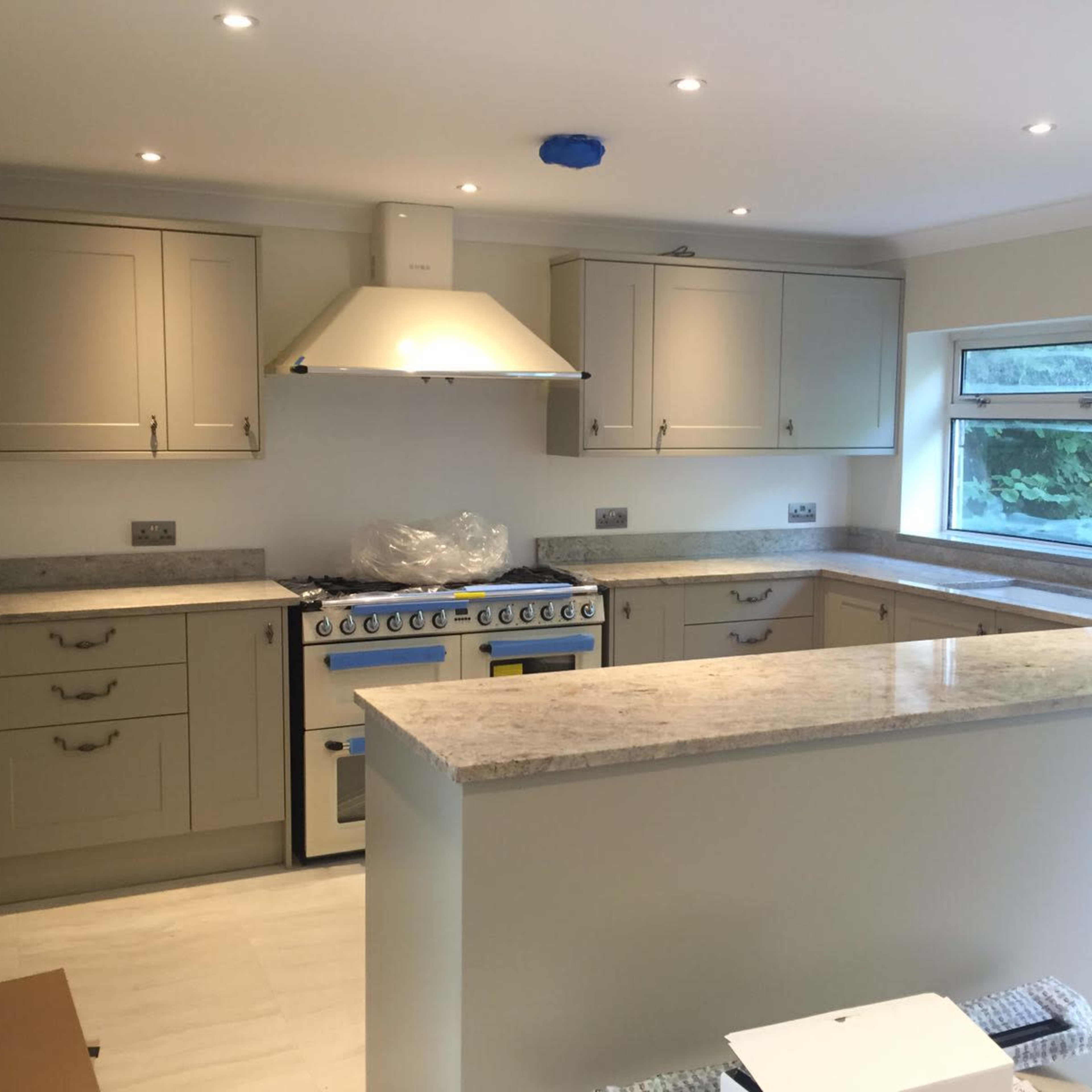 A modern kitchen with light gray cabinetry, a stainless steel range hood, and a central island topped with granite.