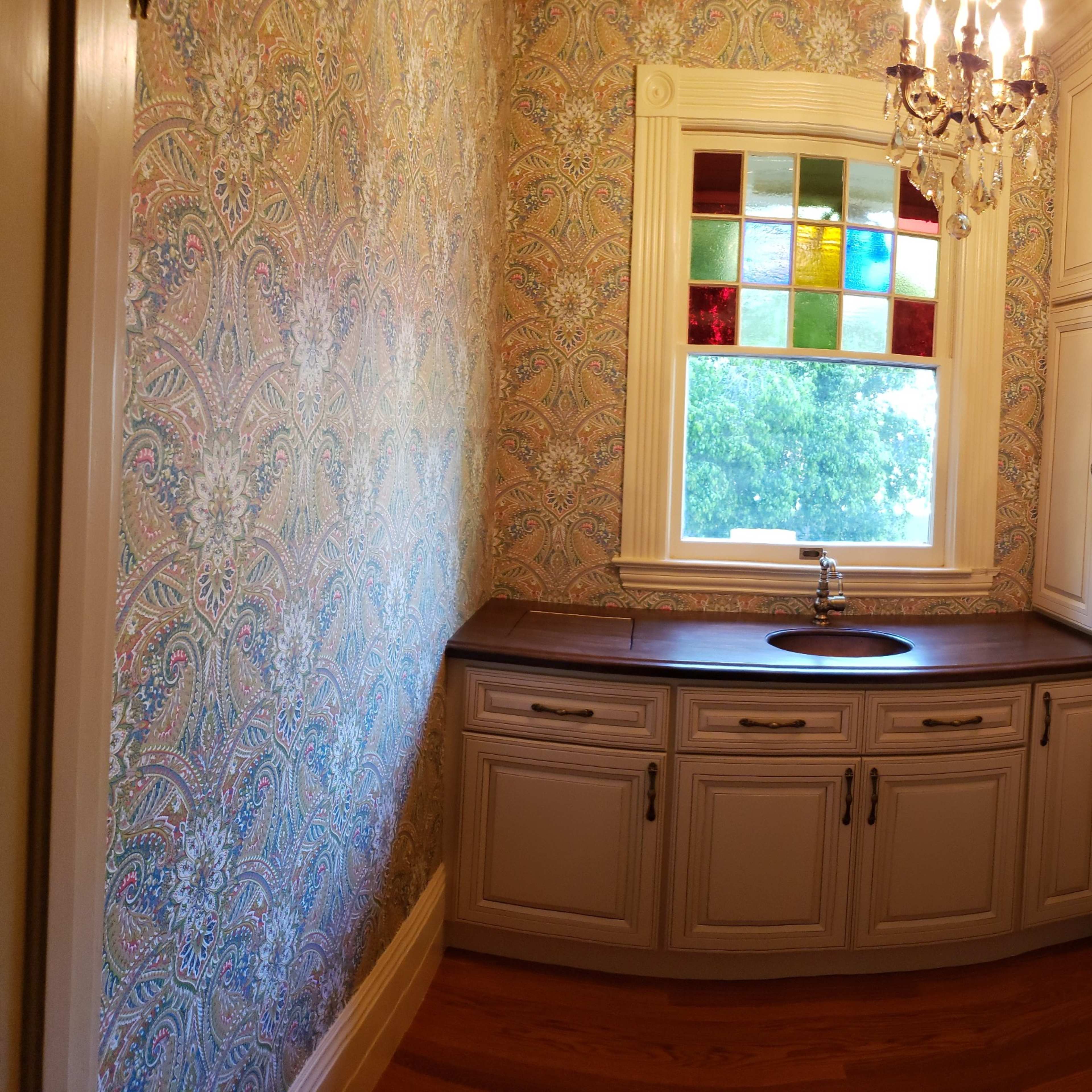 A small room with patterned wallpaper, a wooden countertop, a sink, and a window featuring colored glass panes.
