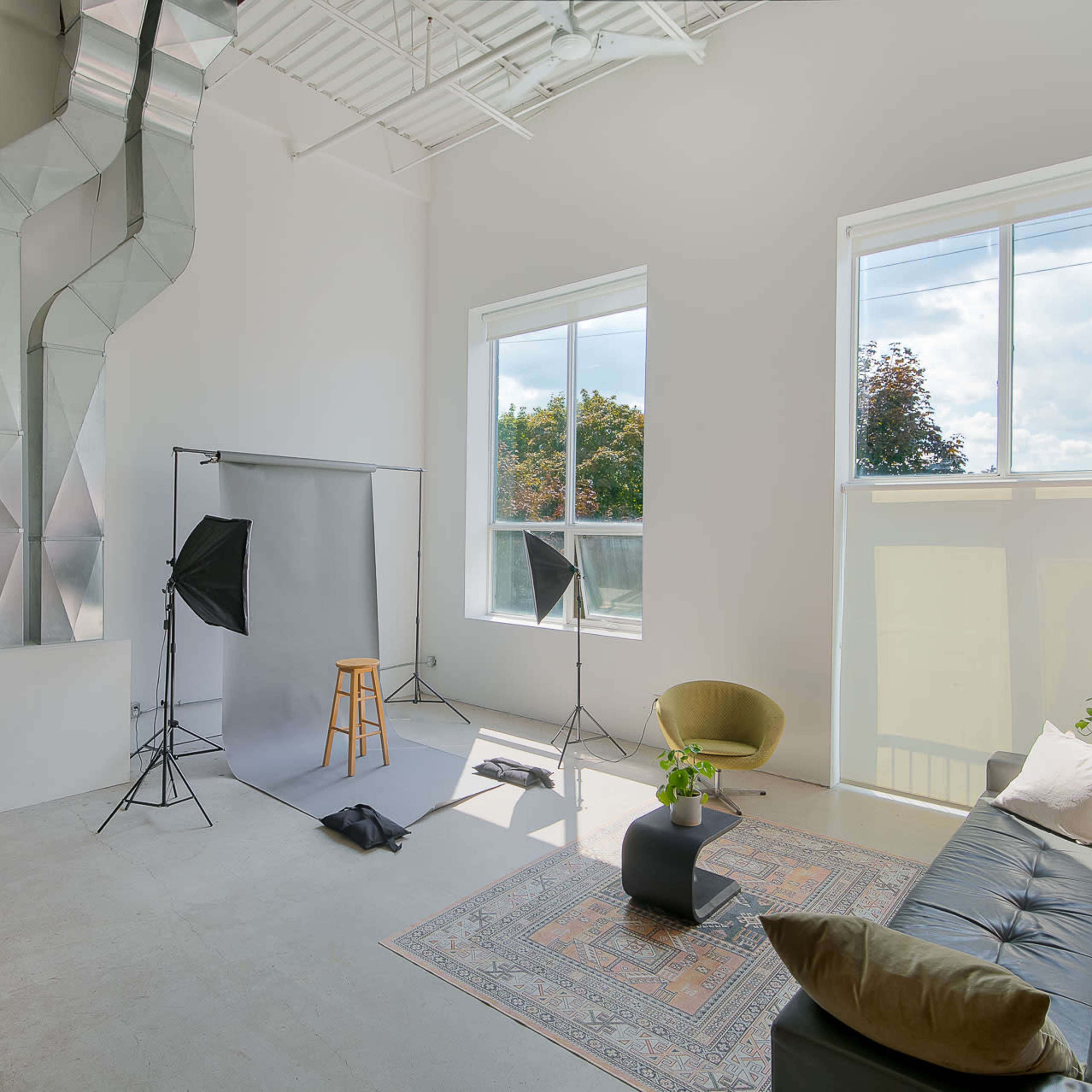 A spacious, well-lit room featuring a large window, a green chair, a dark sofa, and photography equipment set up for a shoot.