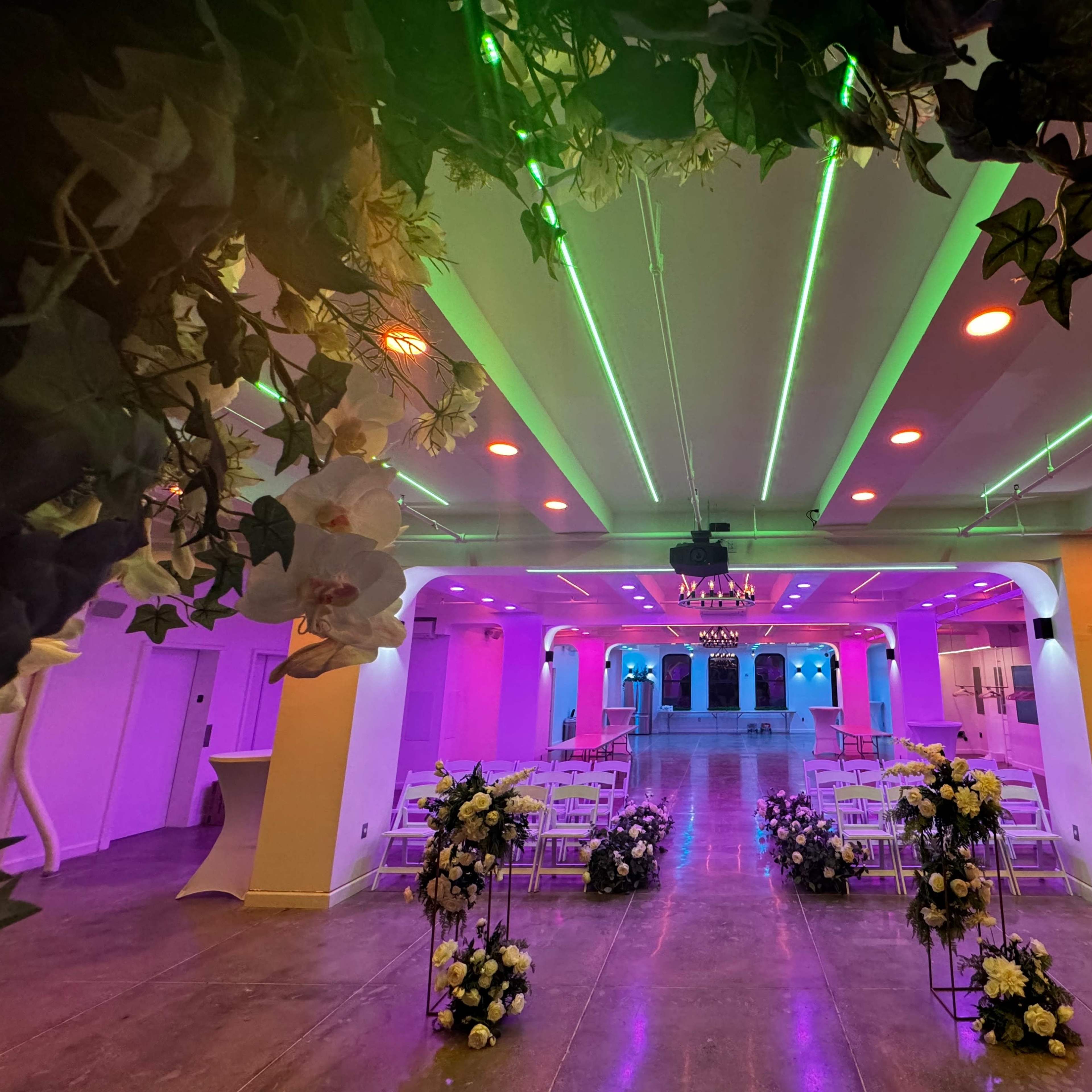 A decorated indoor space features rows of white chairs and floral arrangements, illuminated by colorful lights.