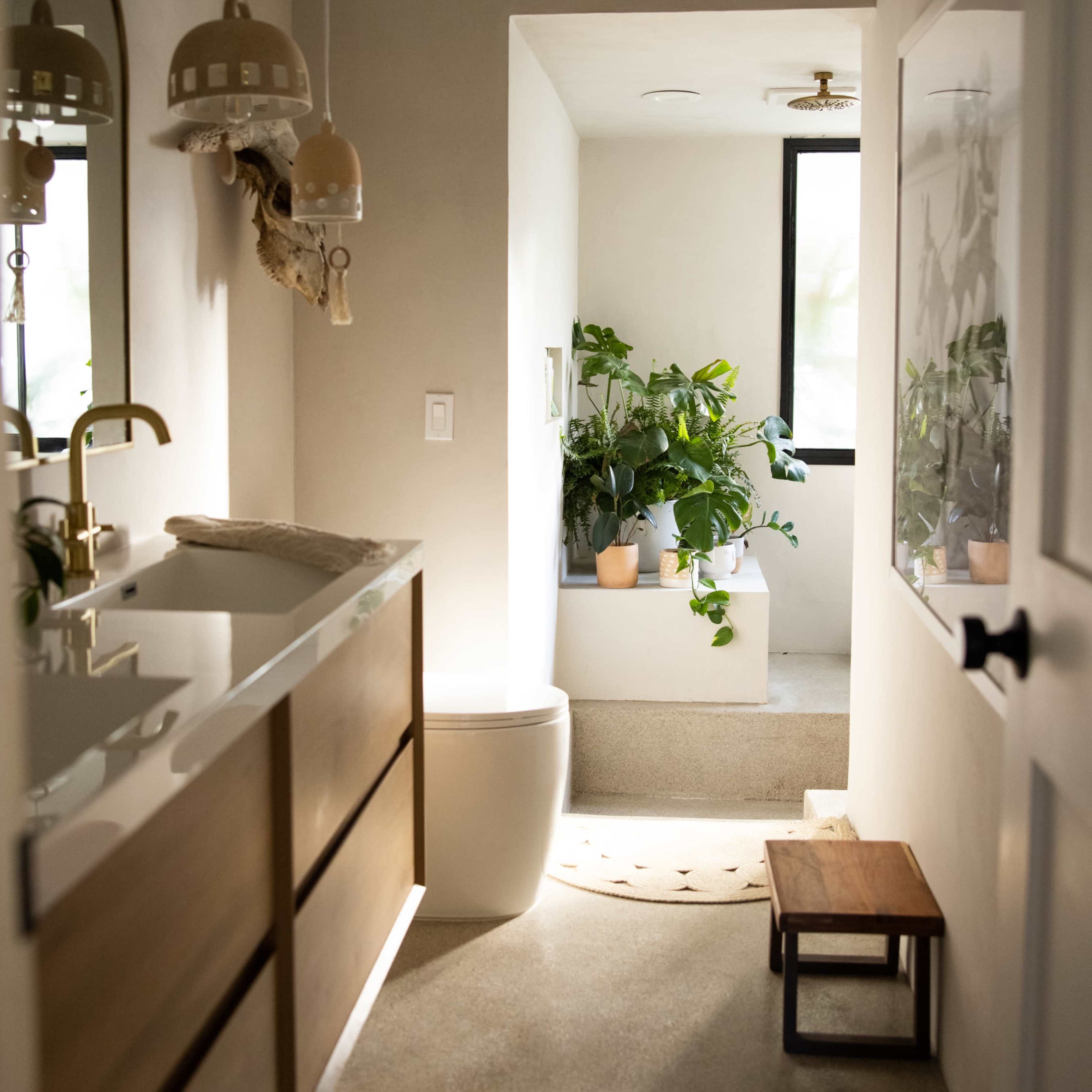 A streamlined bathroom features a wooden vanity, a modern toilet, and a sunlit tub area with potted plants.