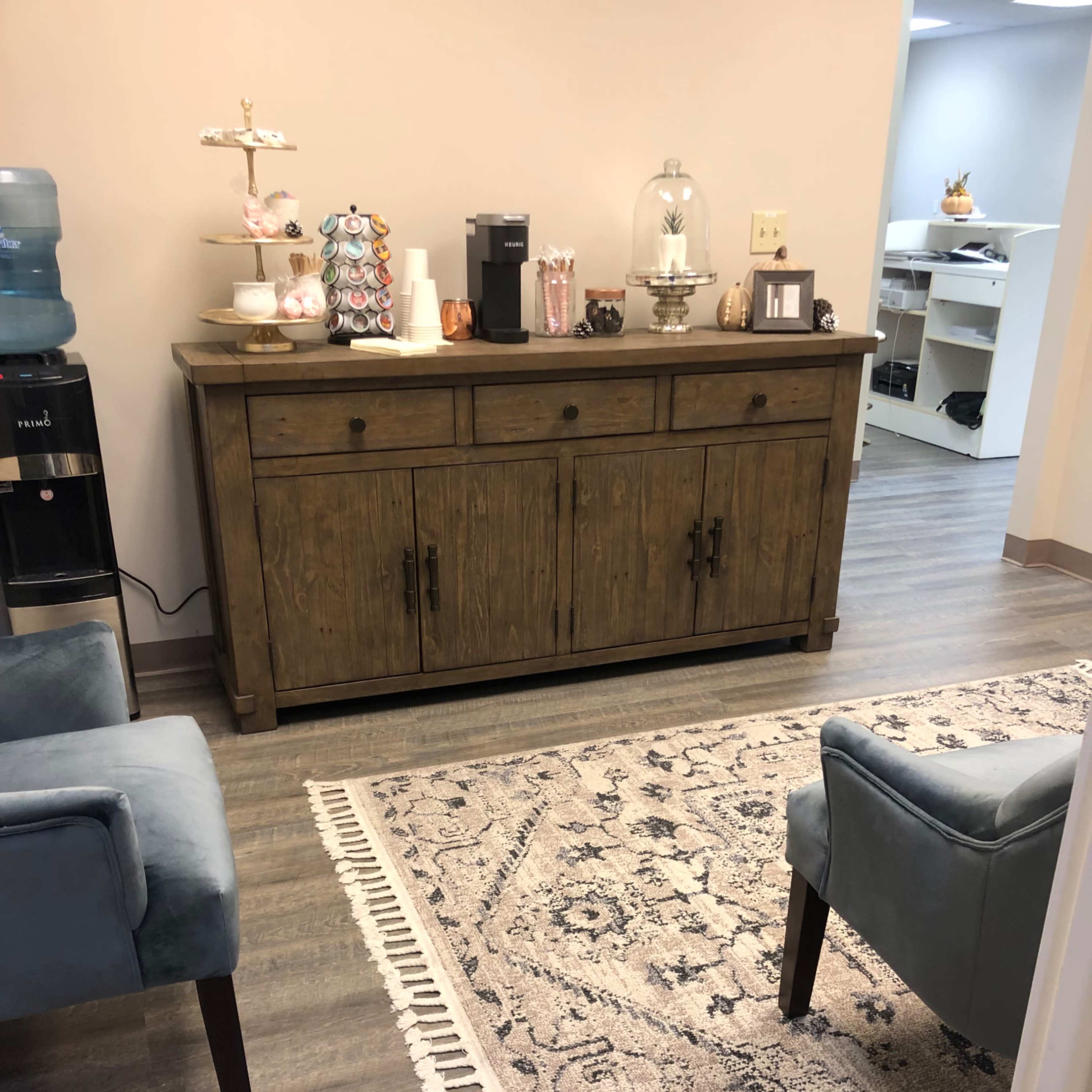 A cozy waiting area with a wooden sideboard and two blue chairs set on a decorative rug.