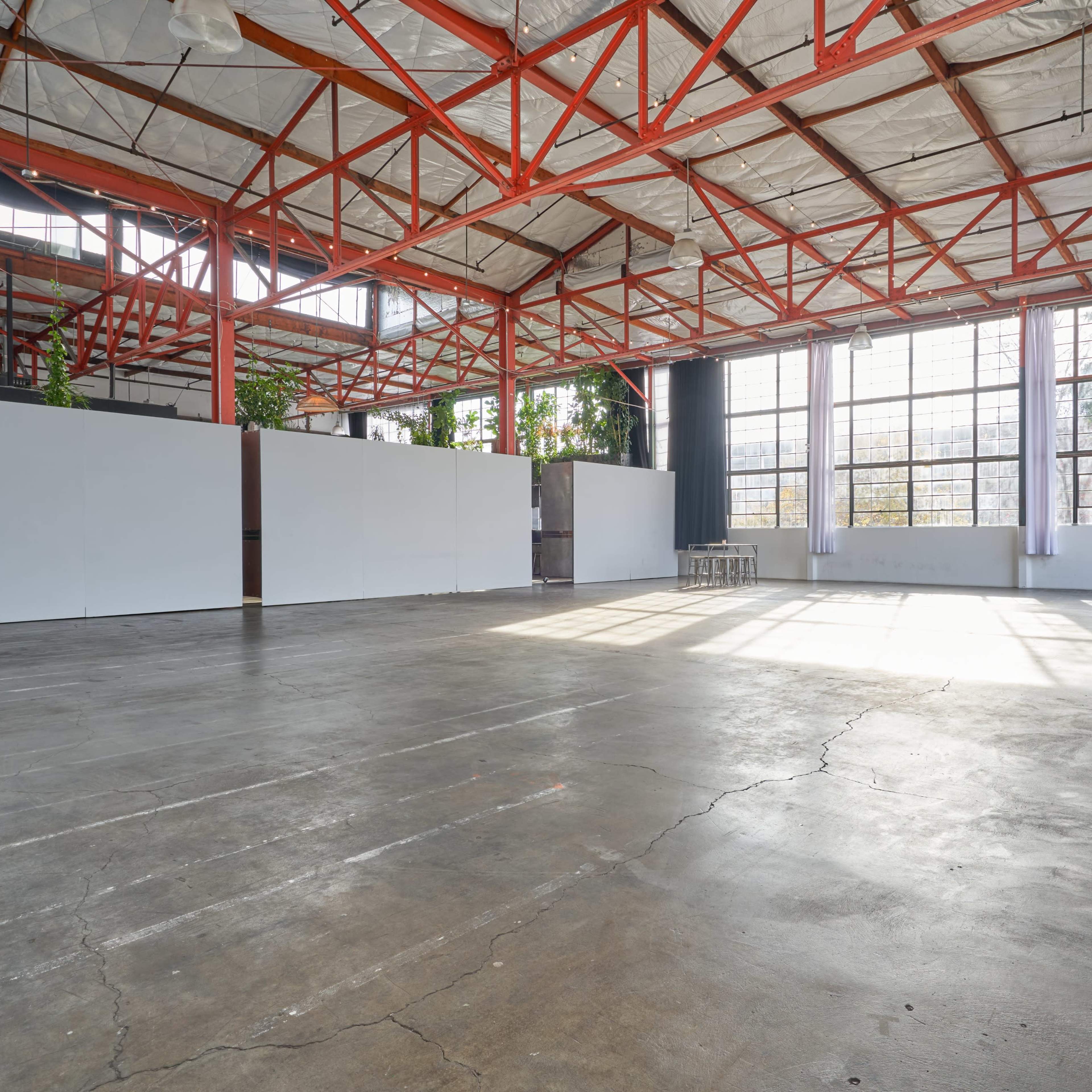 A spacious industrial venue with high ceilings, large windows, and a concrete floor, featuring white wall partitions and greenery.