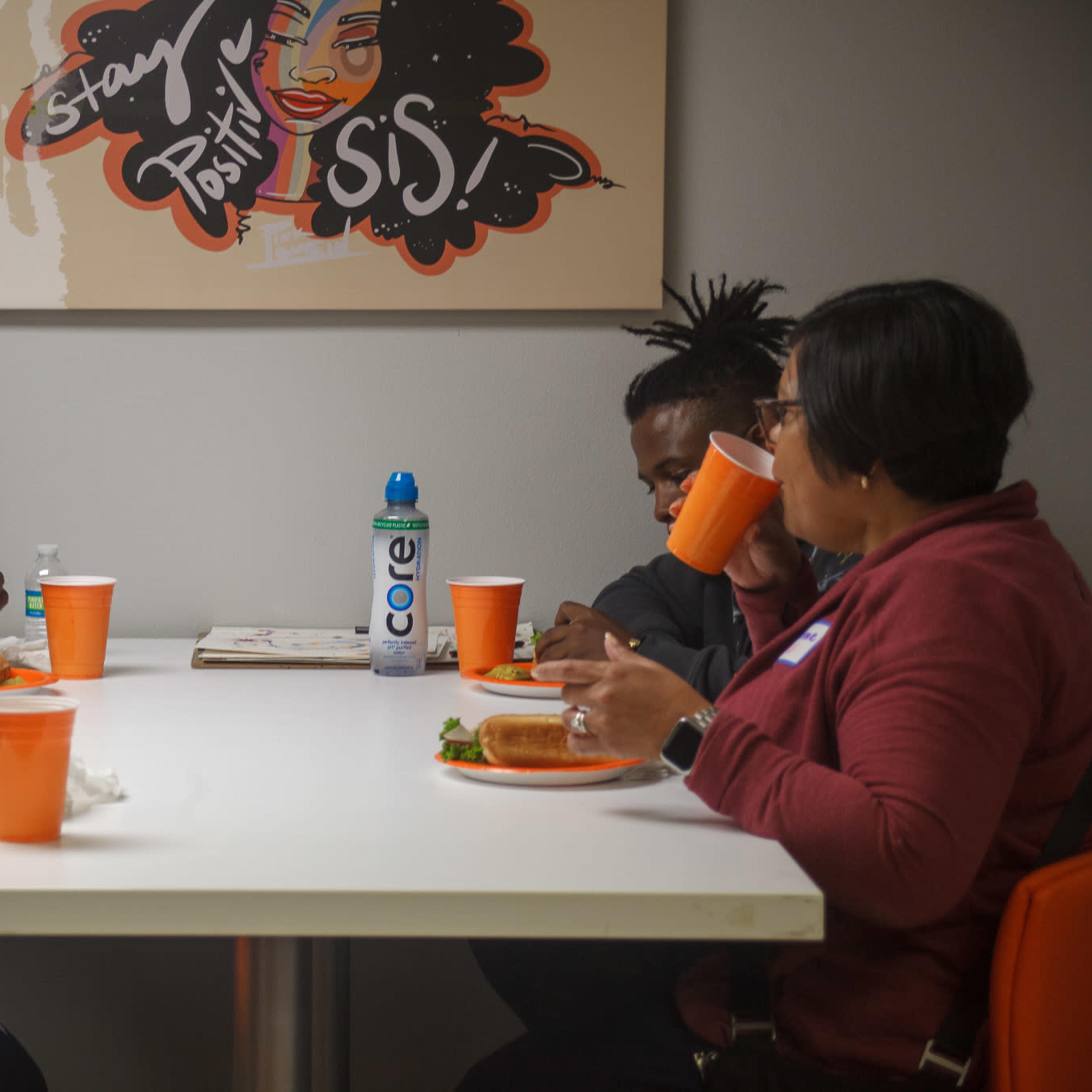 A group of three people sits around a table sharing a meal, with each person holding an orange cup in a brightly colored room.