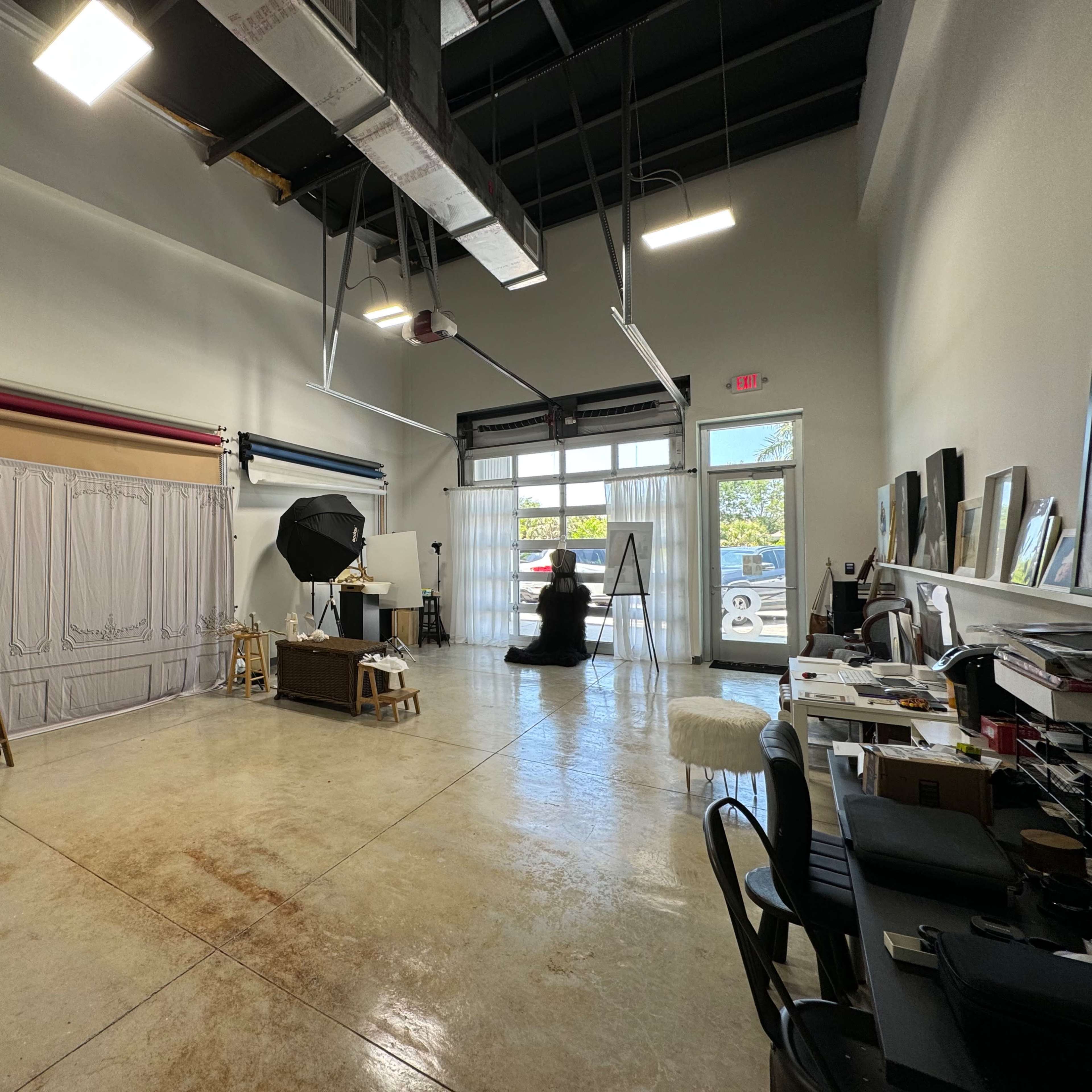 A spacious studio with a backdrop setup, a large light umbrella, various furniture pieces, and a view of the outside through glass doors.