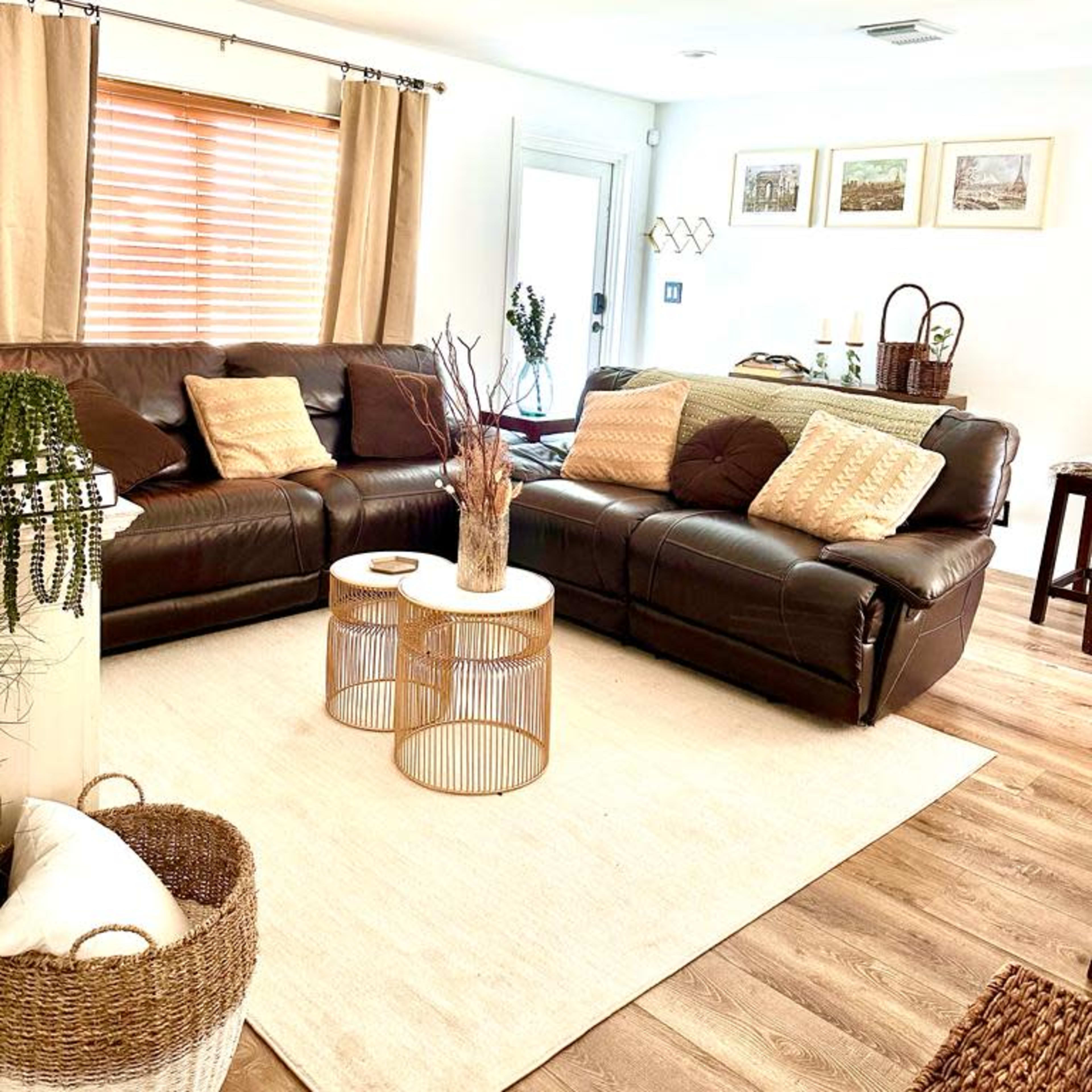 The living room features a brown leather sectional sofa, a round coffee table, and neutral-colored decor with window treatments and wall art.