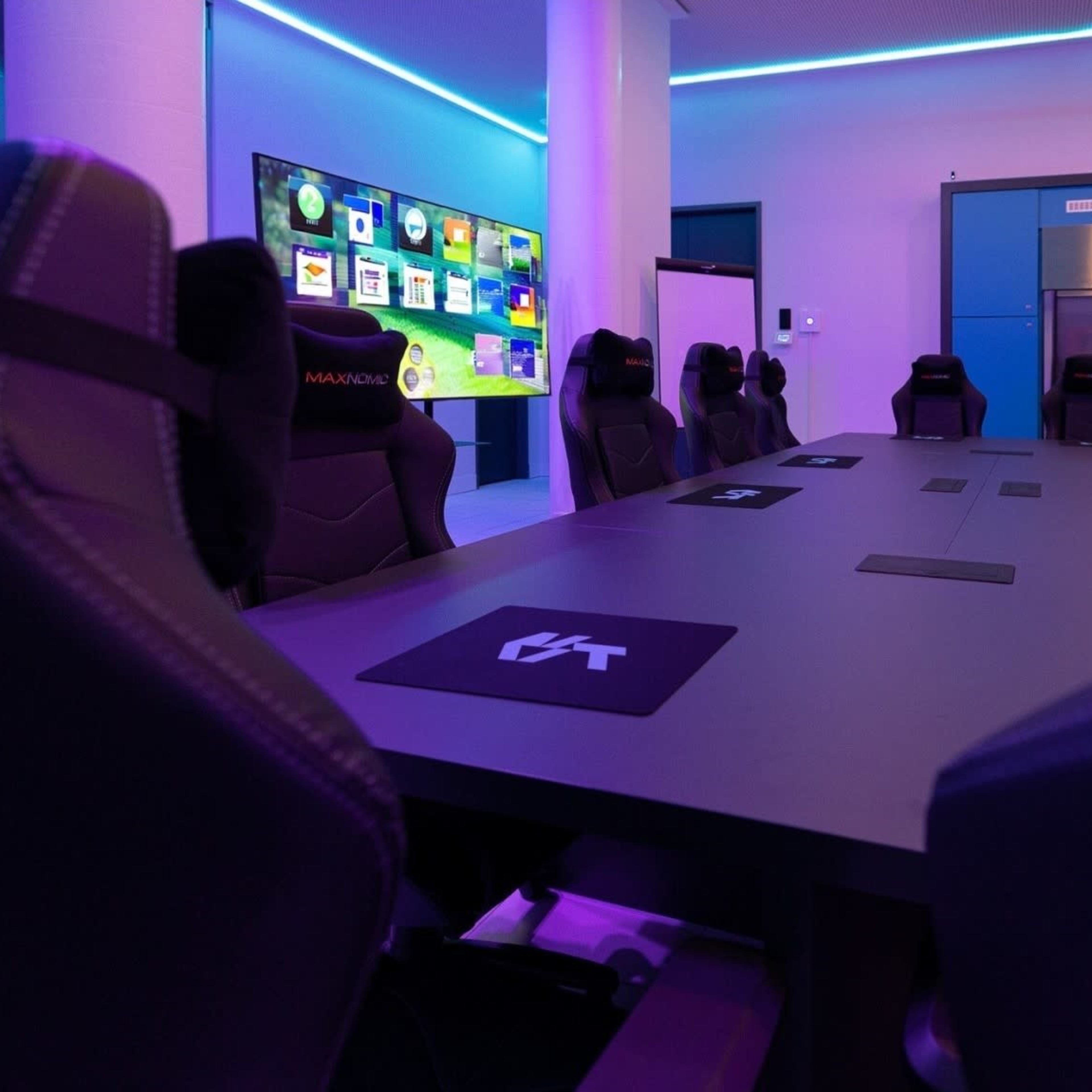 A modern conference room features a long table surrounded by ergonomic chairs, with a large screen displaying colorful graphics on the wall.