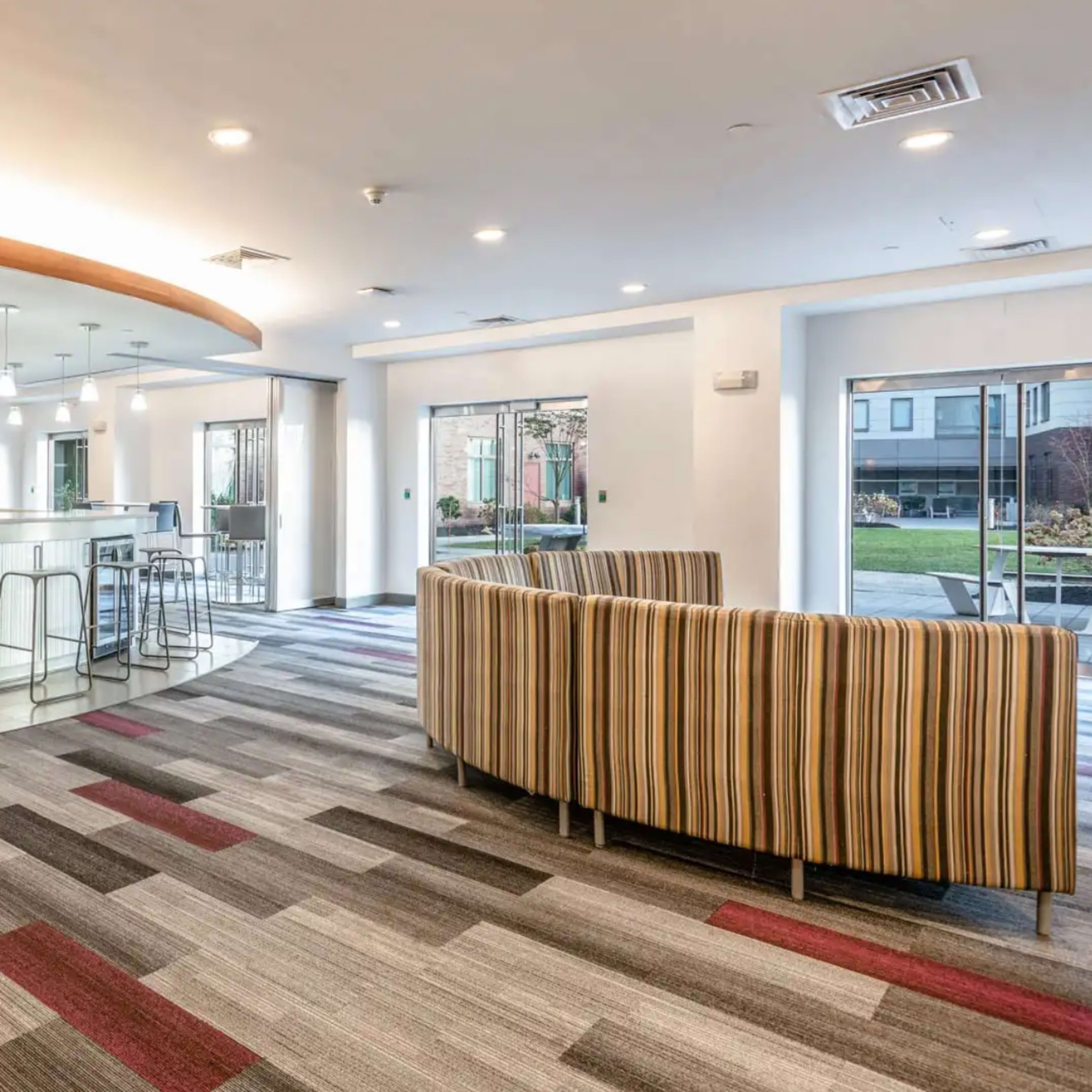 A modern common area with a striped sofa, a bar-style counter, and a dining area with high stools, all situated on patterned carpet.