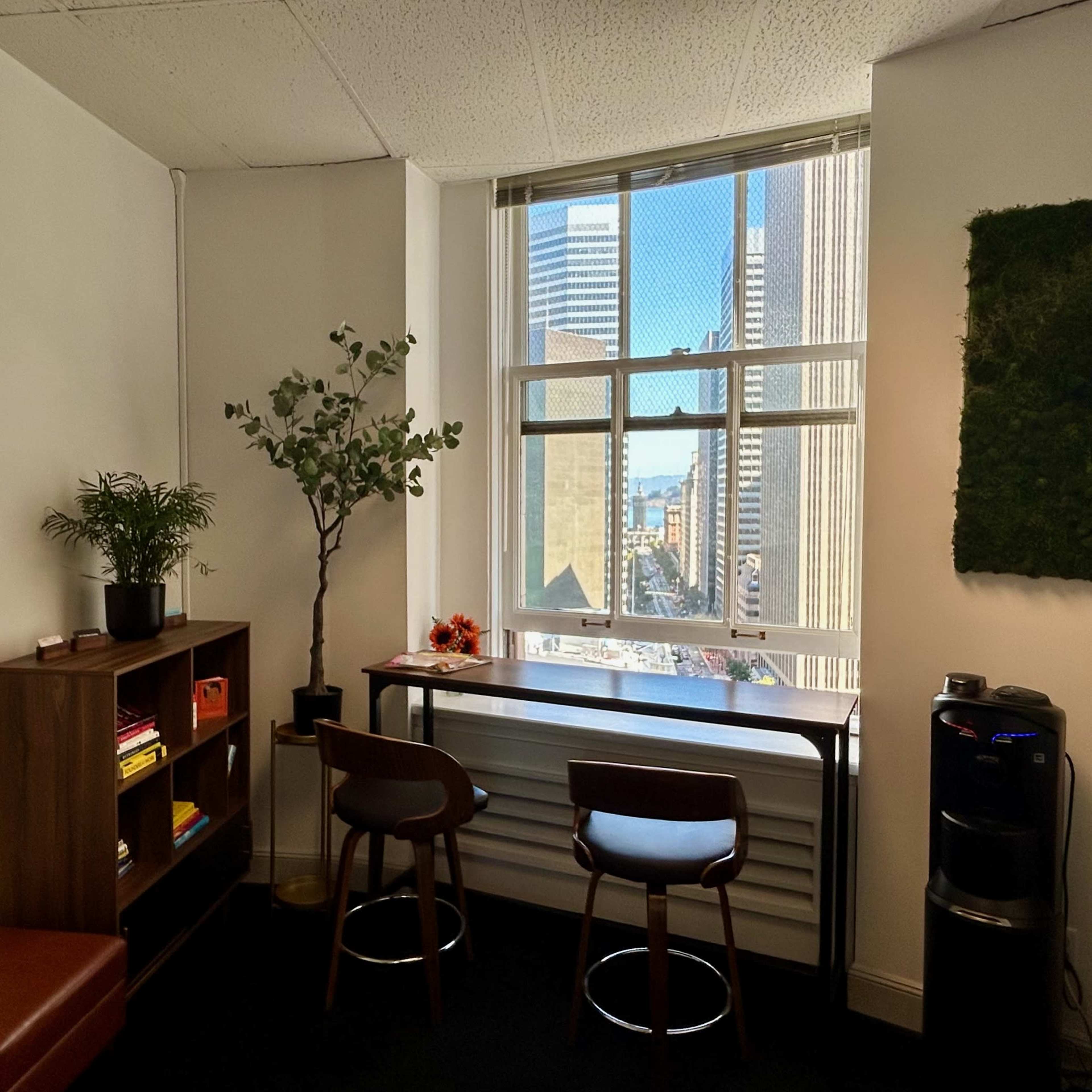 An office space with a window overlooking a cityscape, featuring a small table with two chairs and a plant nearby.