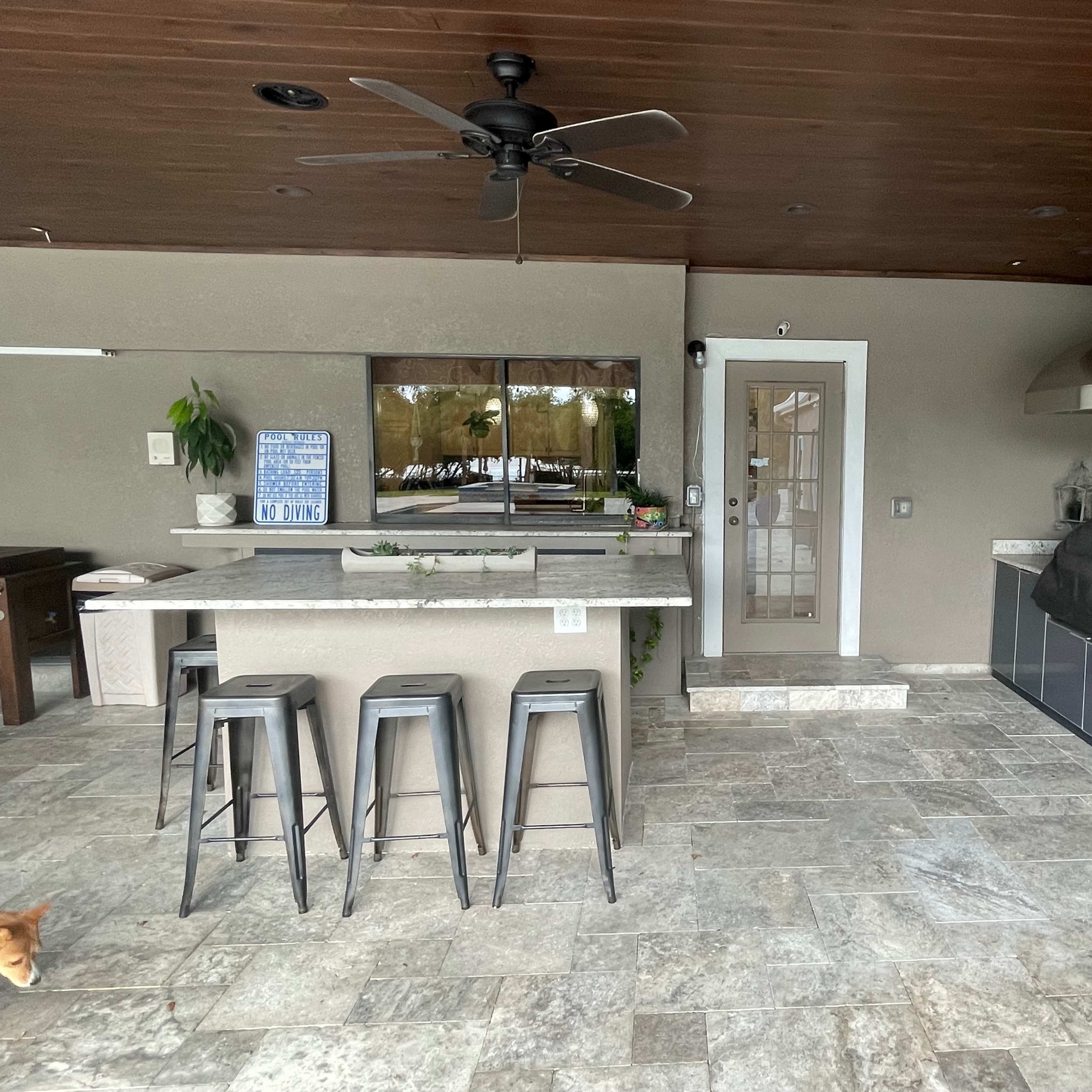 A covered outdoor kitchen area featuring a stone floor, an island with stools, and stainless steel appliances.