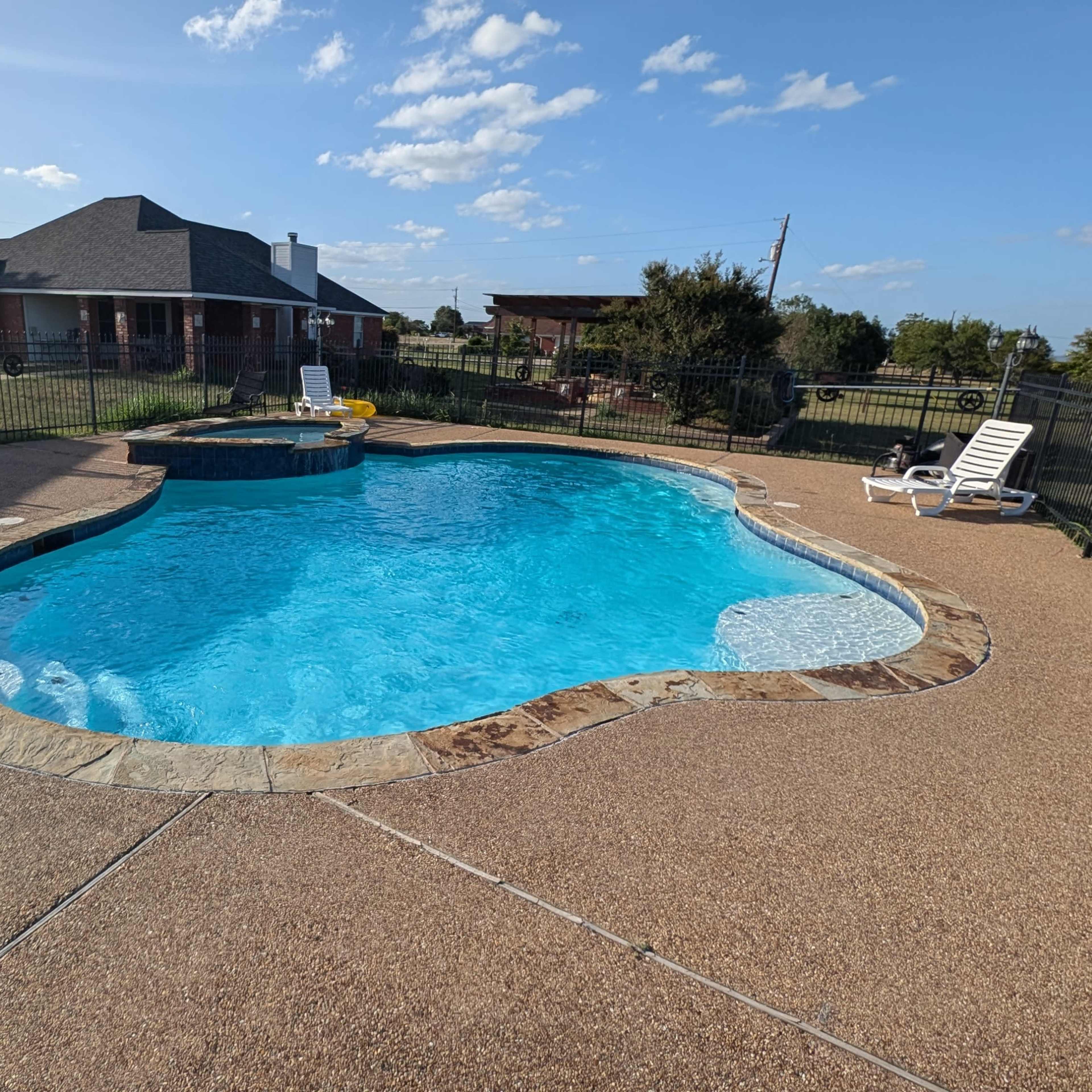 A clear blue pool with a unique shape is surrounded by a stone deck and a fenced area, with a house and grassy landscape visible in the background.