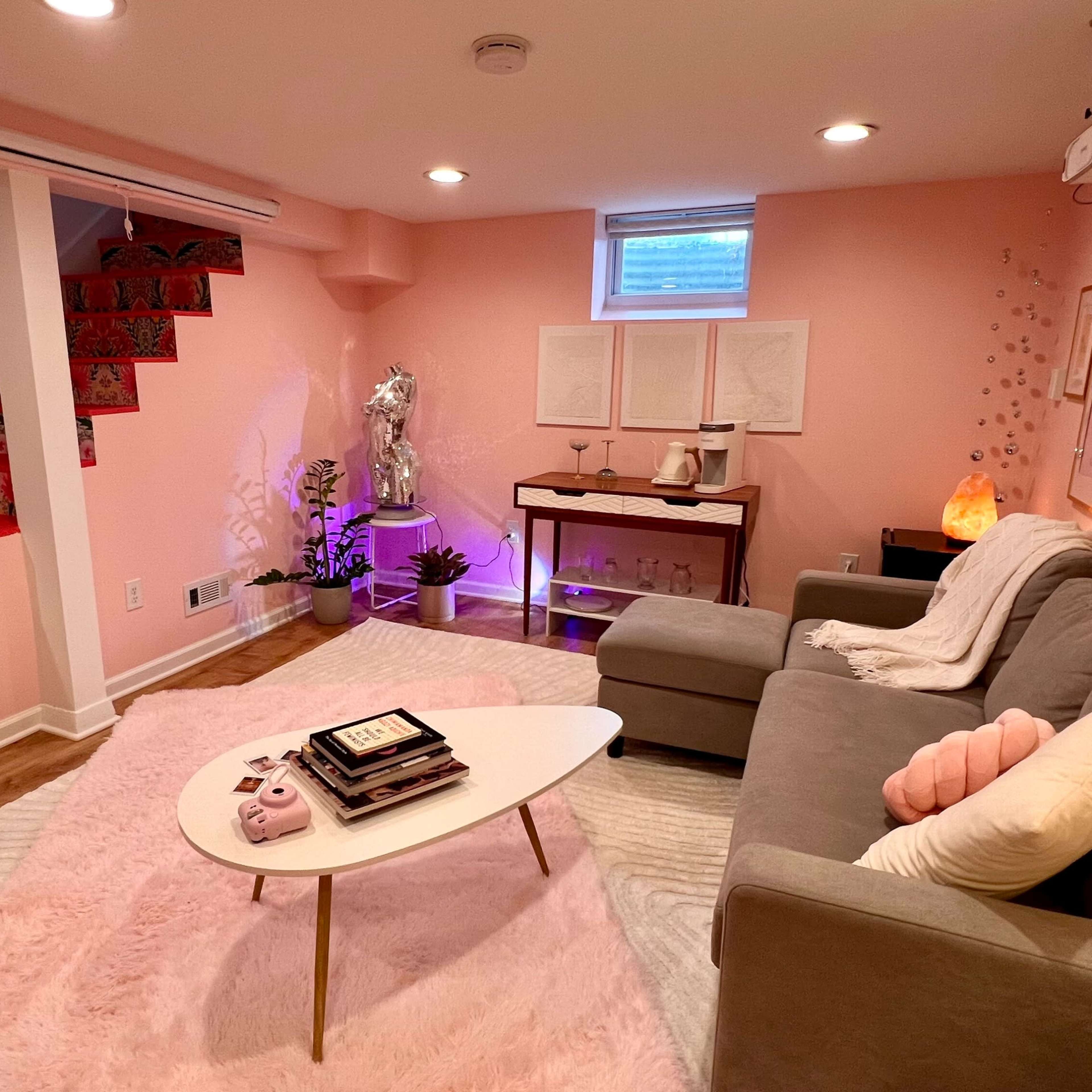 A cozy basement living area with a pink color scheme, featuring a gray sofa, a coffee table, decorative art on the walls, and a staircase with patterned steps.