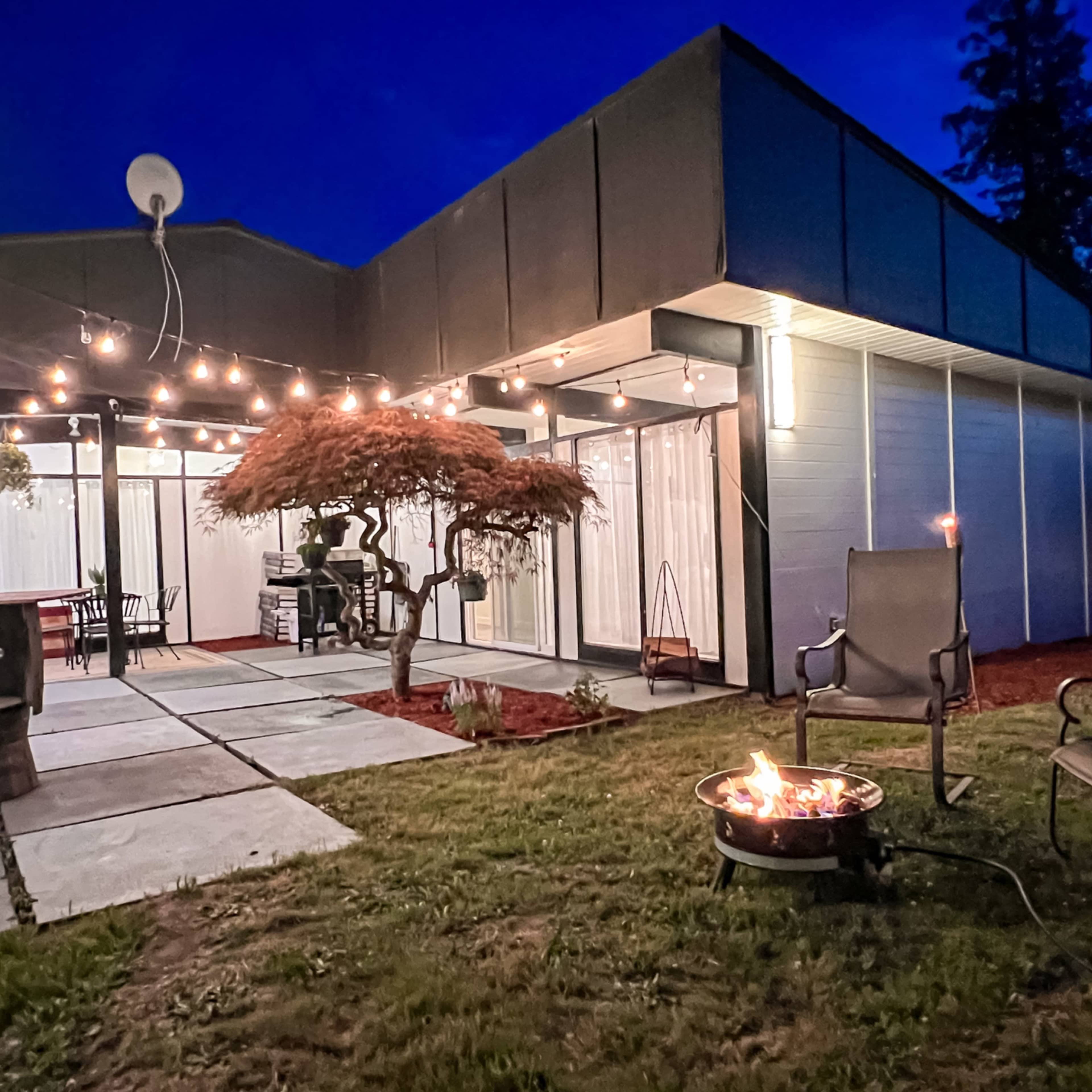 A backyard patio features stone pavers, string lights overhead, a fire pit, and a small tree in the garden area.