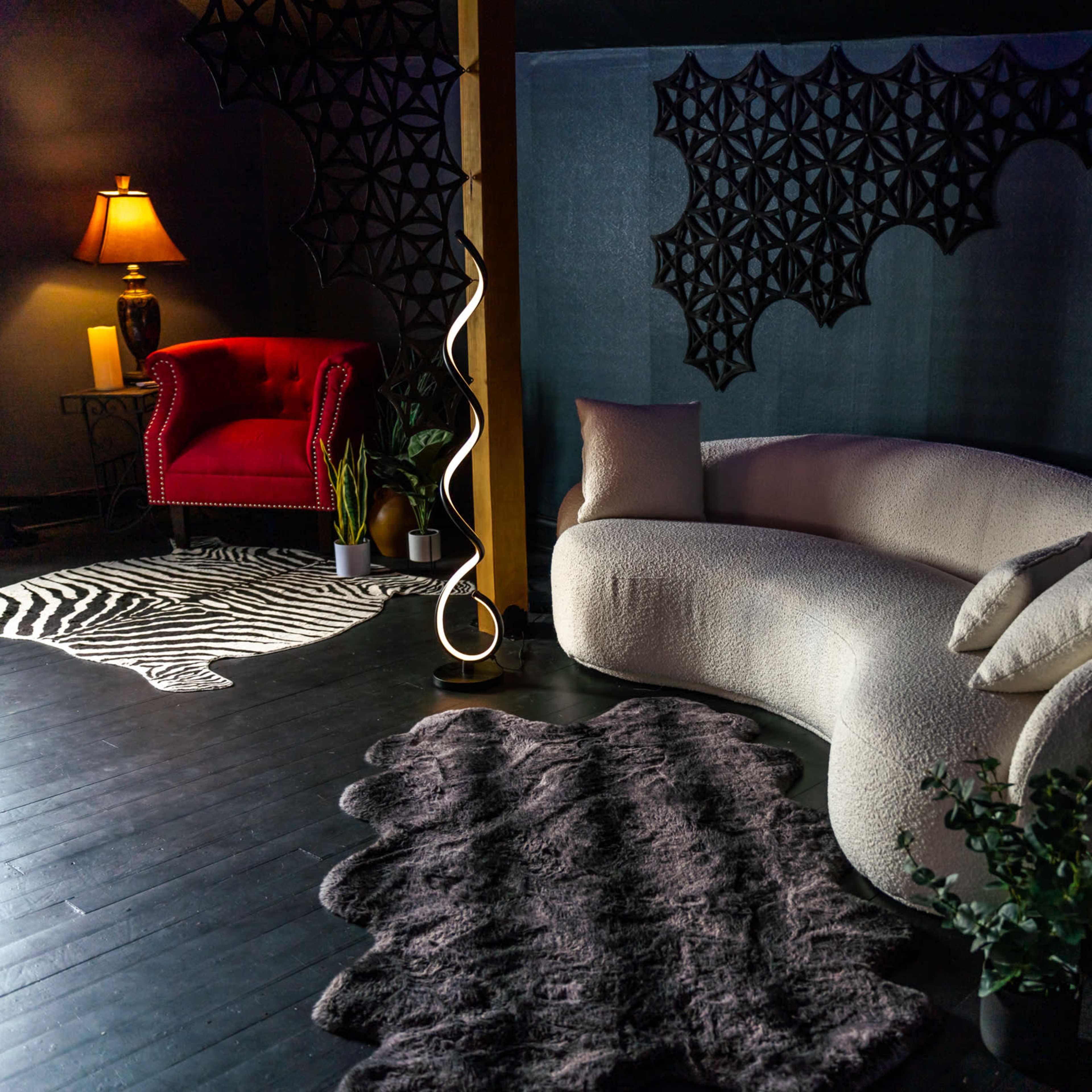 A modern living space featuring a curved white sofa, a red armchair, decorative plants, and various textured rugs against a dark backdrop.