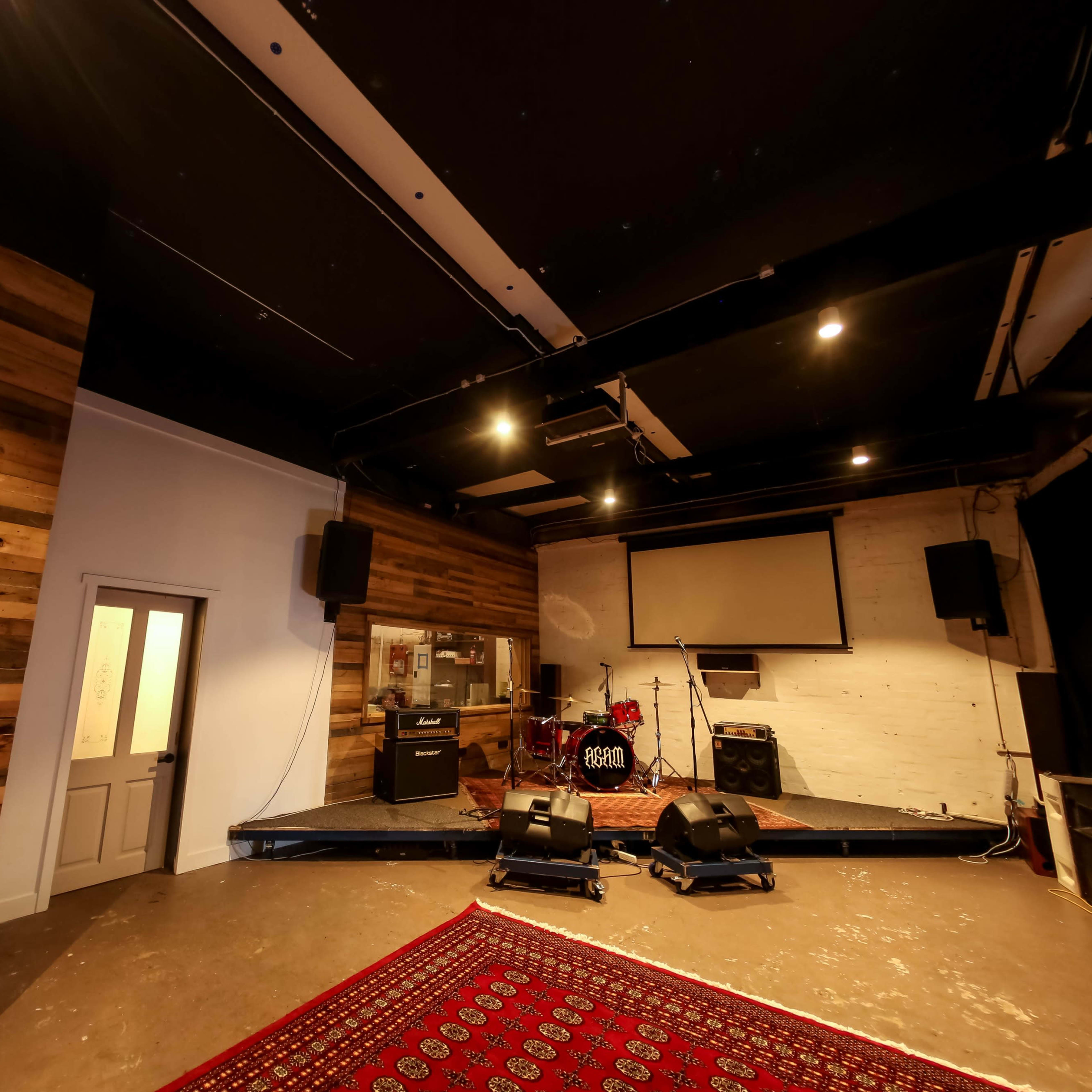 A music performance space with a wooden wall, a small stage, audio equipment, and a red patterned rug on the floor.