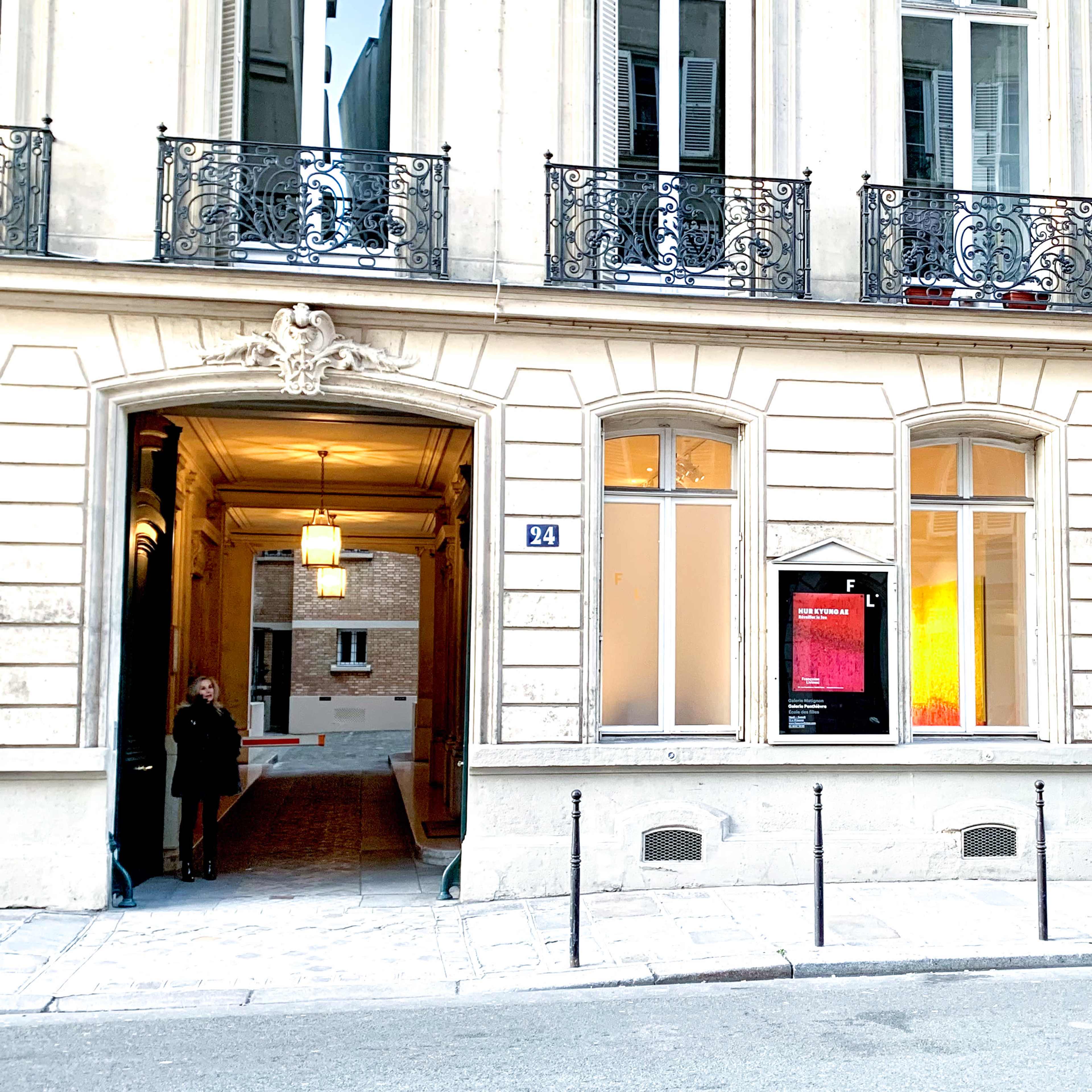 A person stands outside a building with tall windows and intricate ironwork, located on a quiet street.