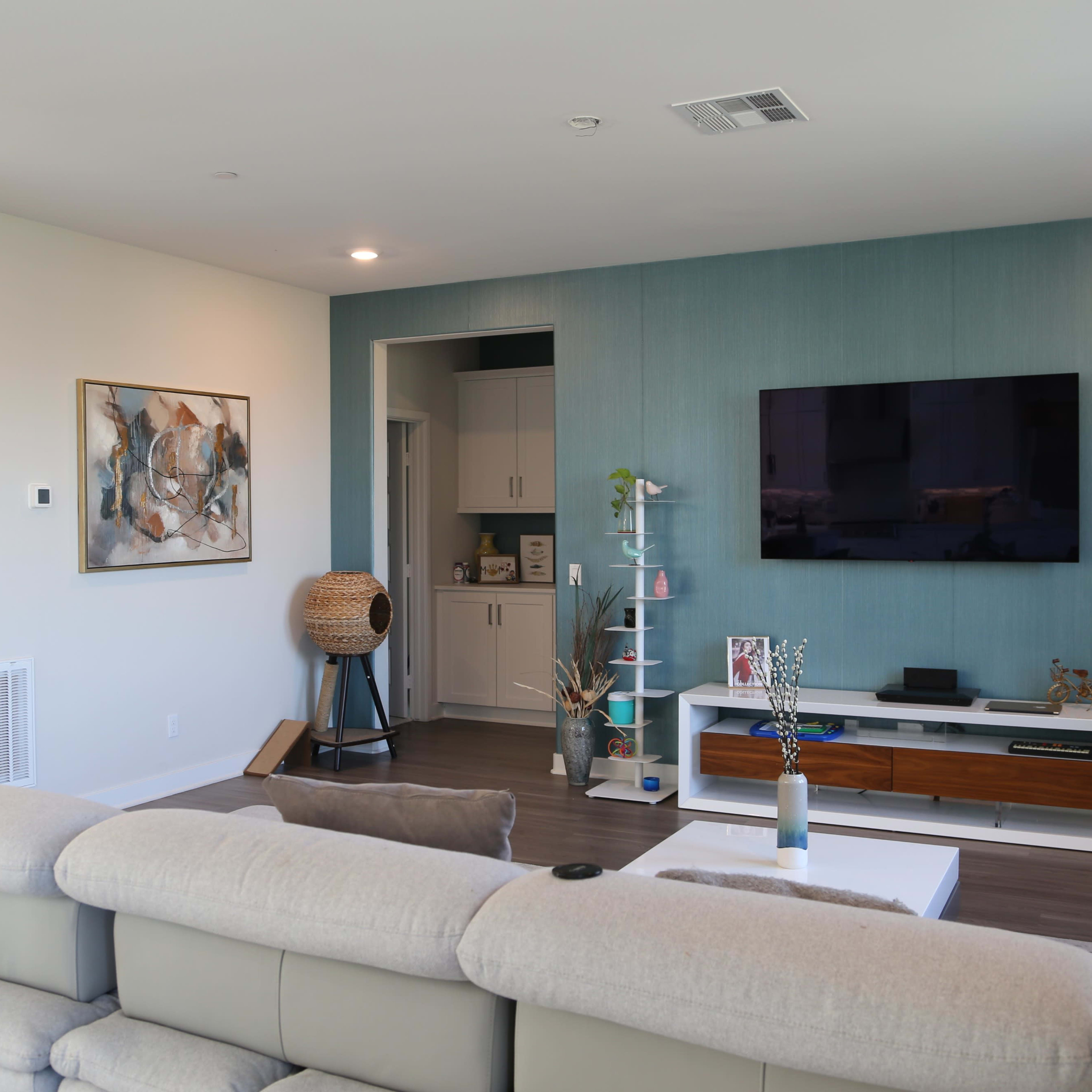 A modern living room features a light-colored sectional sofa, a large wall-mounted TV, and a coffee table, with decorative elements including a painting and a plant.