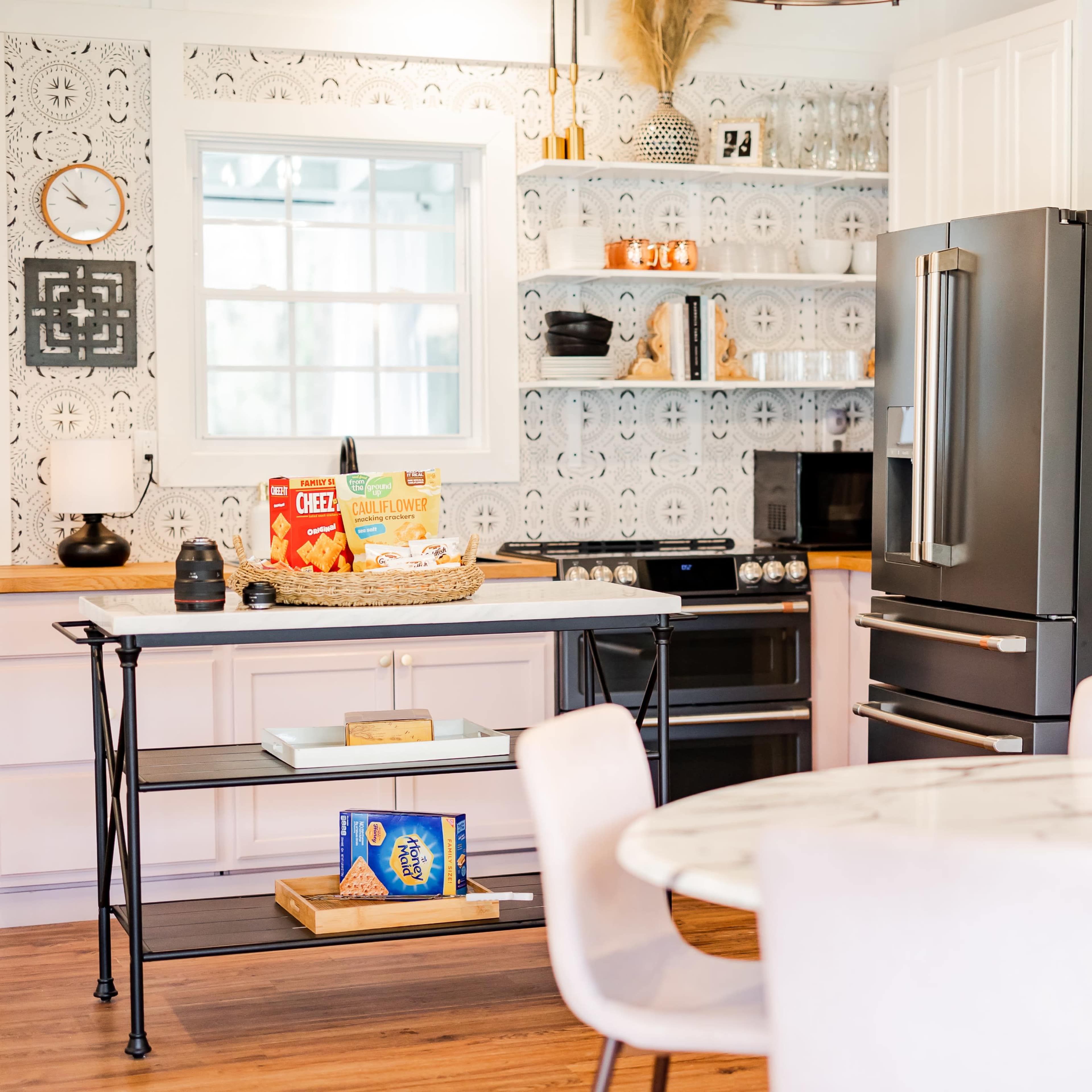 The kitchen features a black refrigerator, white cabinetry, and a round marble dining table, with decorative elements on the walls and a vase of flowers on the counter.