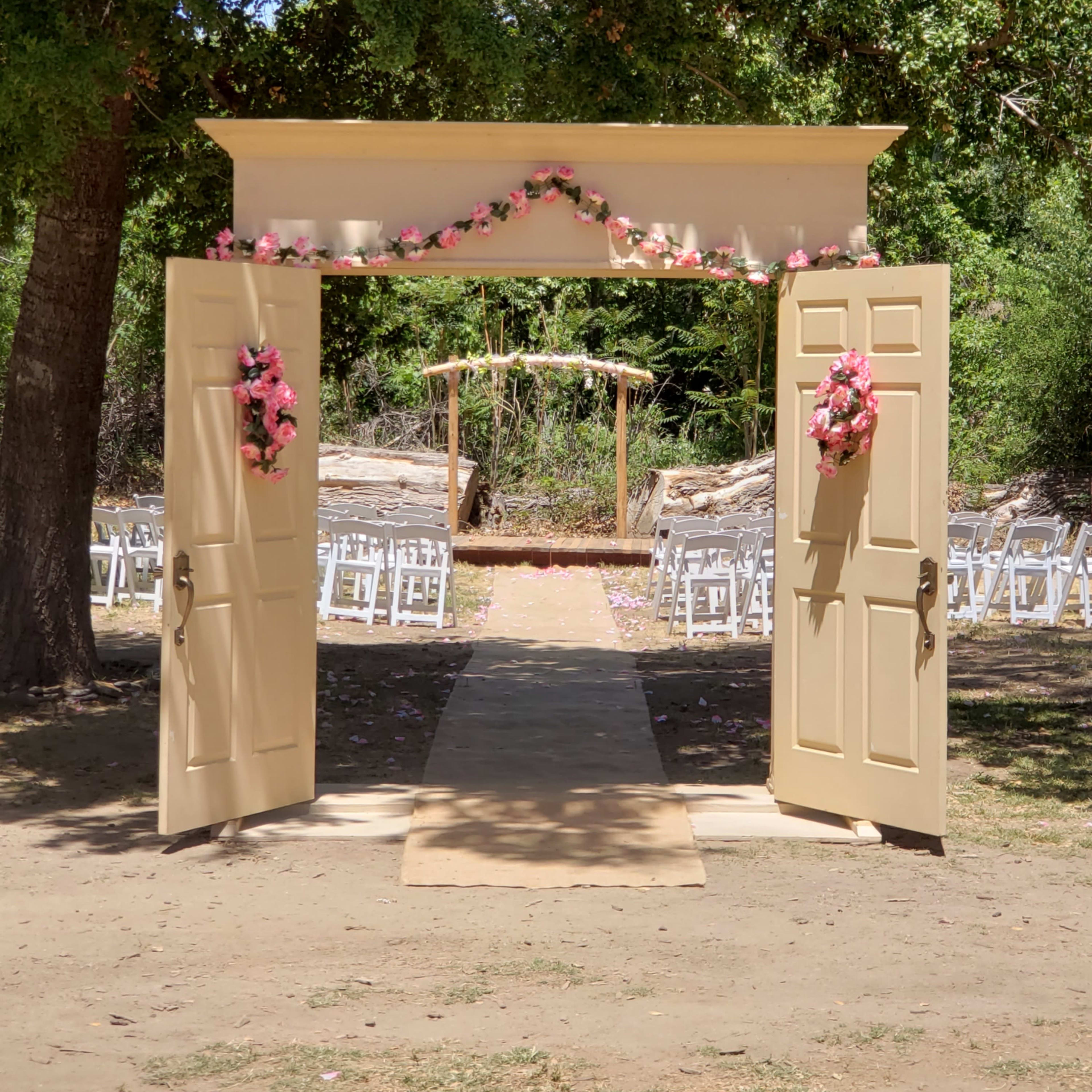 An open double door adorned with pink flowers leads to an outdoor wedding ceremony space featuring chairs and a decorative arch.