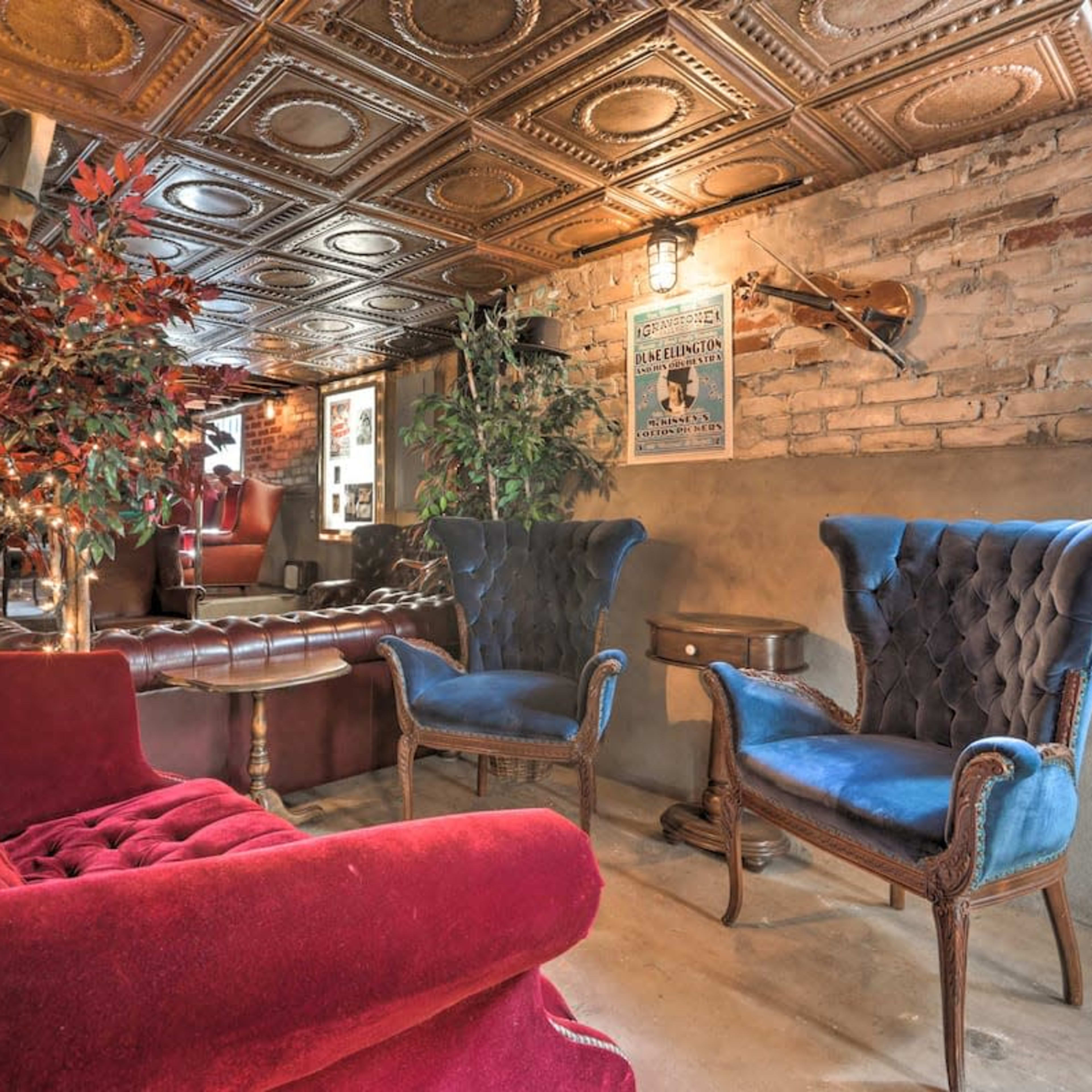 A cozy lounge area featuring a red velvet sofa and two blue armchairs, alongside exposed brick walls and patterned tin ceilings.