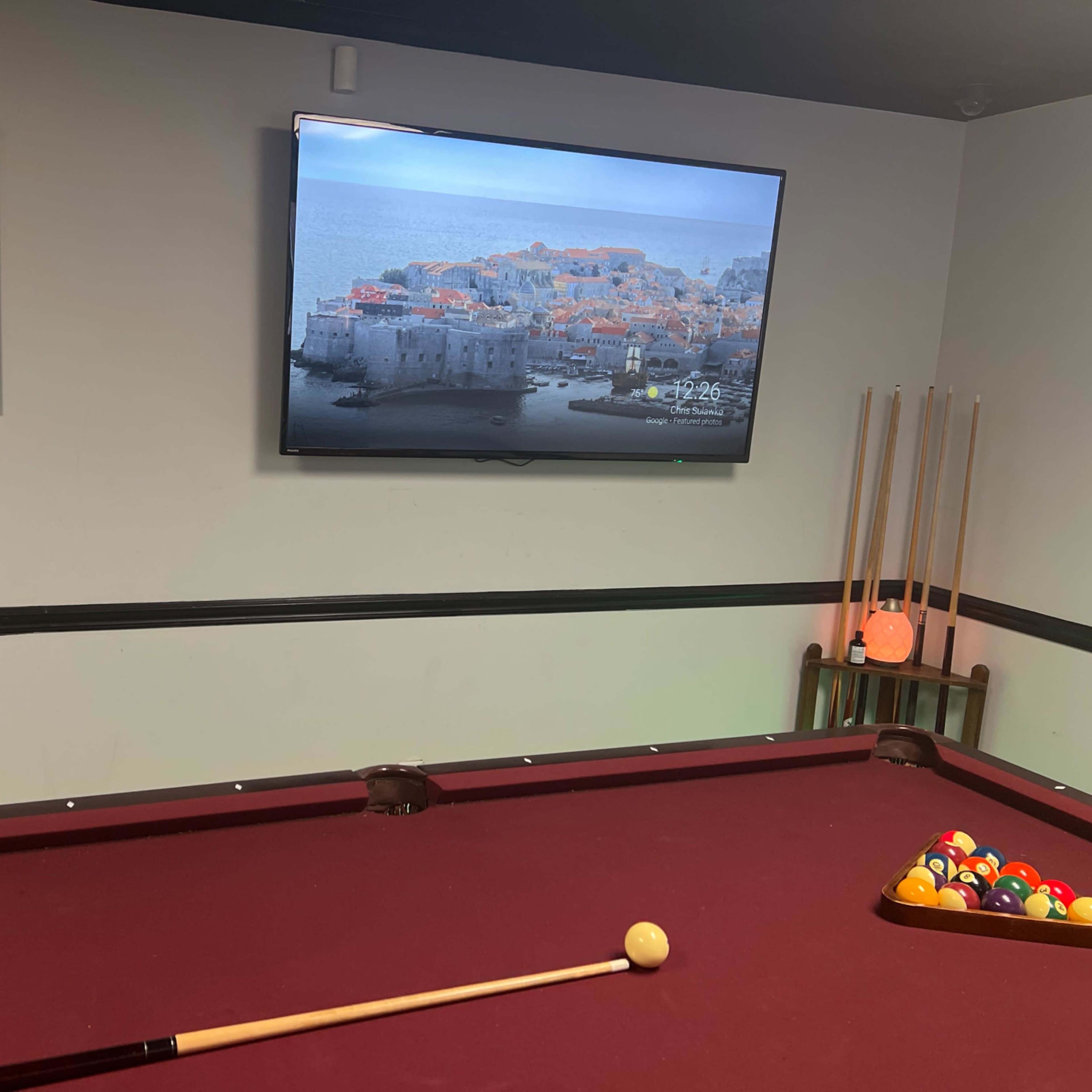 A game room with a red pool table, a television displaying a coastal scene, and a wall art piece that says "FOLLOW YOUR DREAMS."