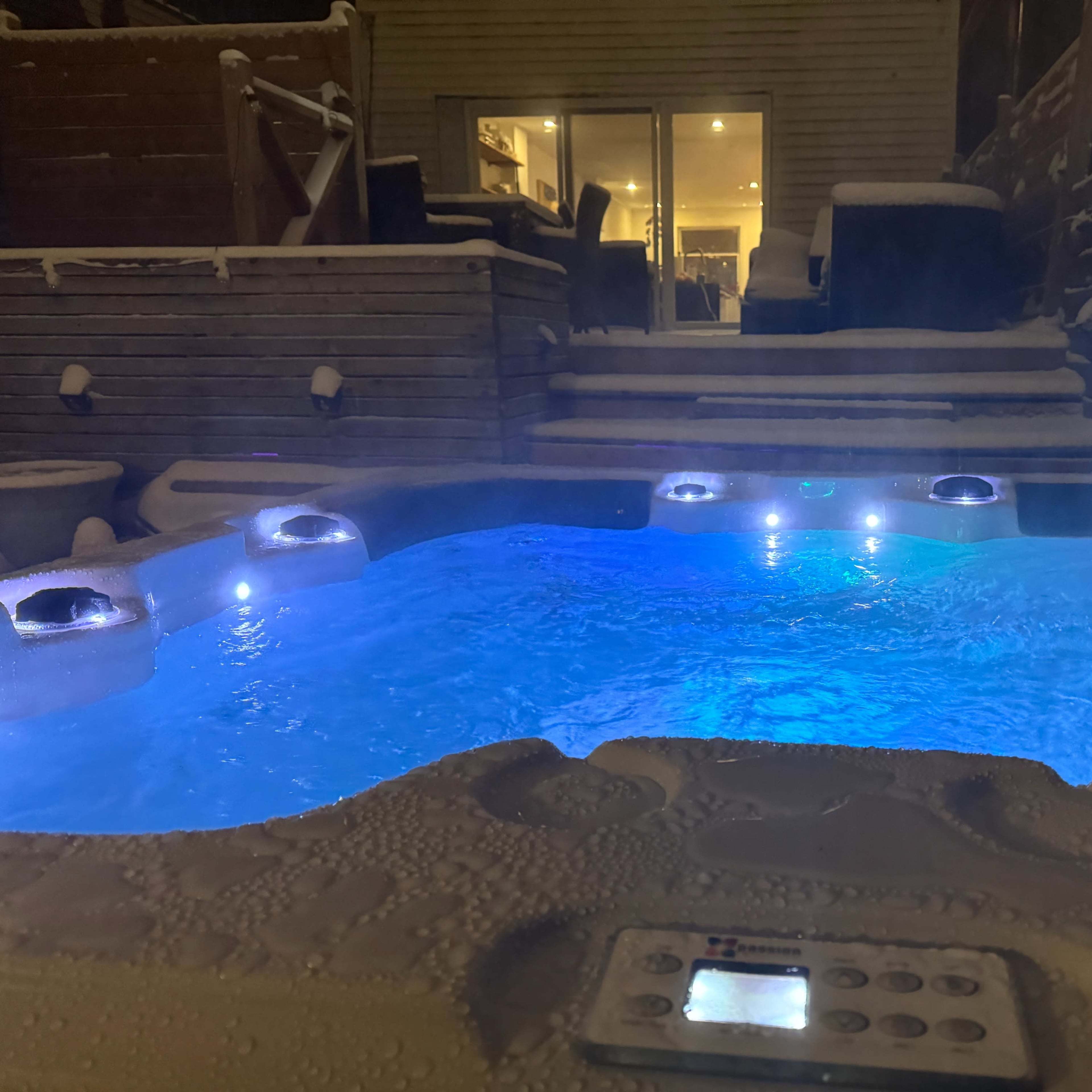 A hot tub filled with blue water and illuminated by underwater lights is positioned on a snowy terrace near a wooden deck and large glass doors.