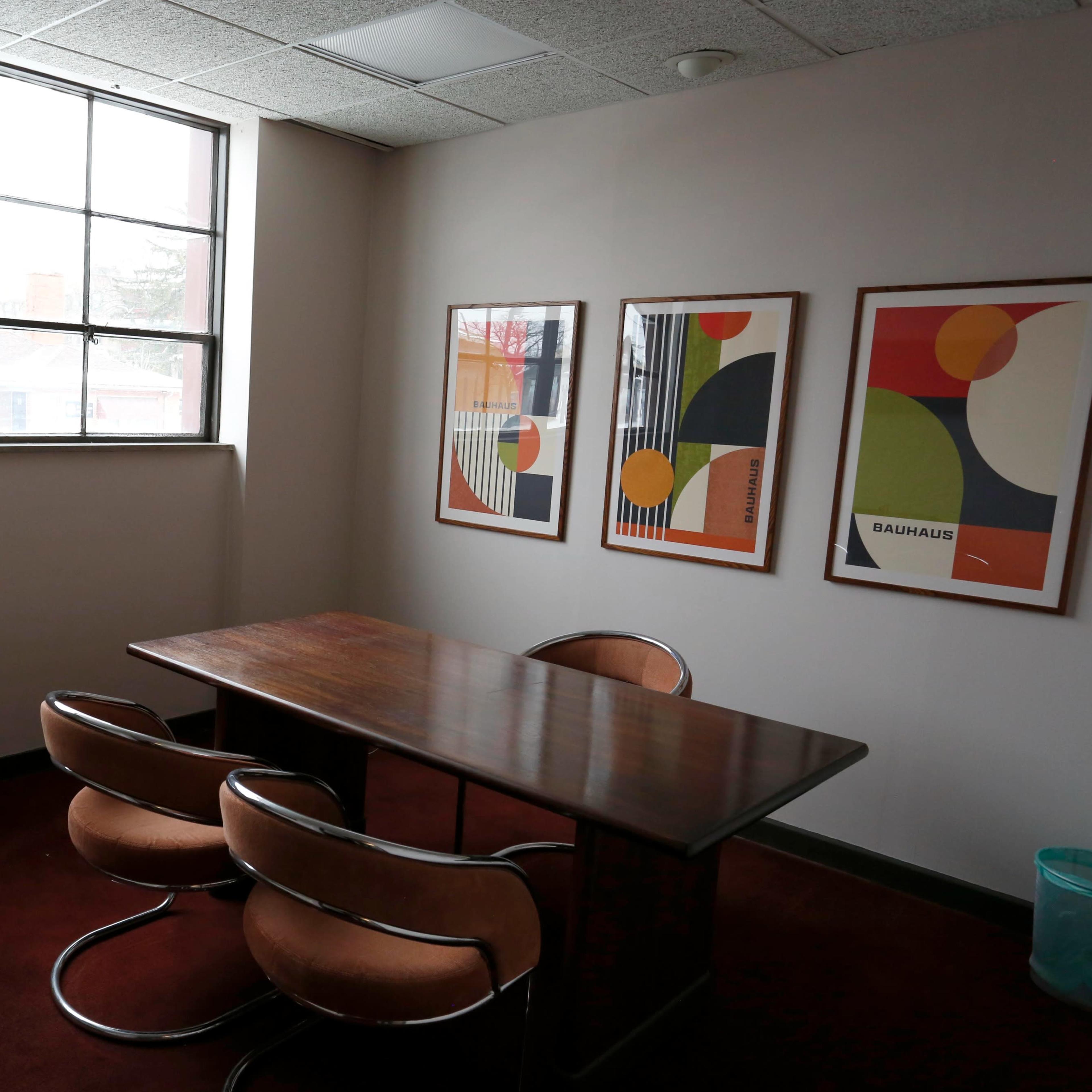 A small office features a wooden table with three circular chairs and three framed abstract art pieces on the walls.