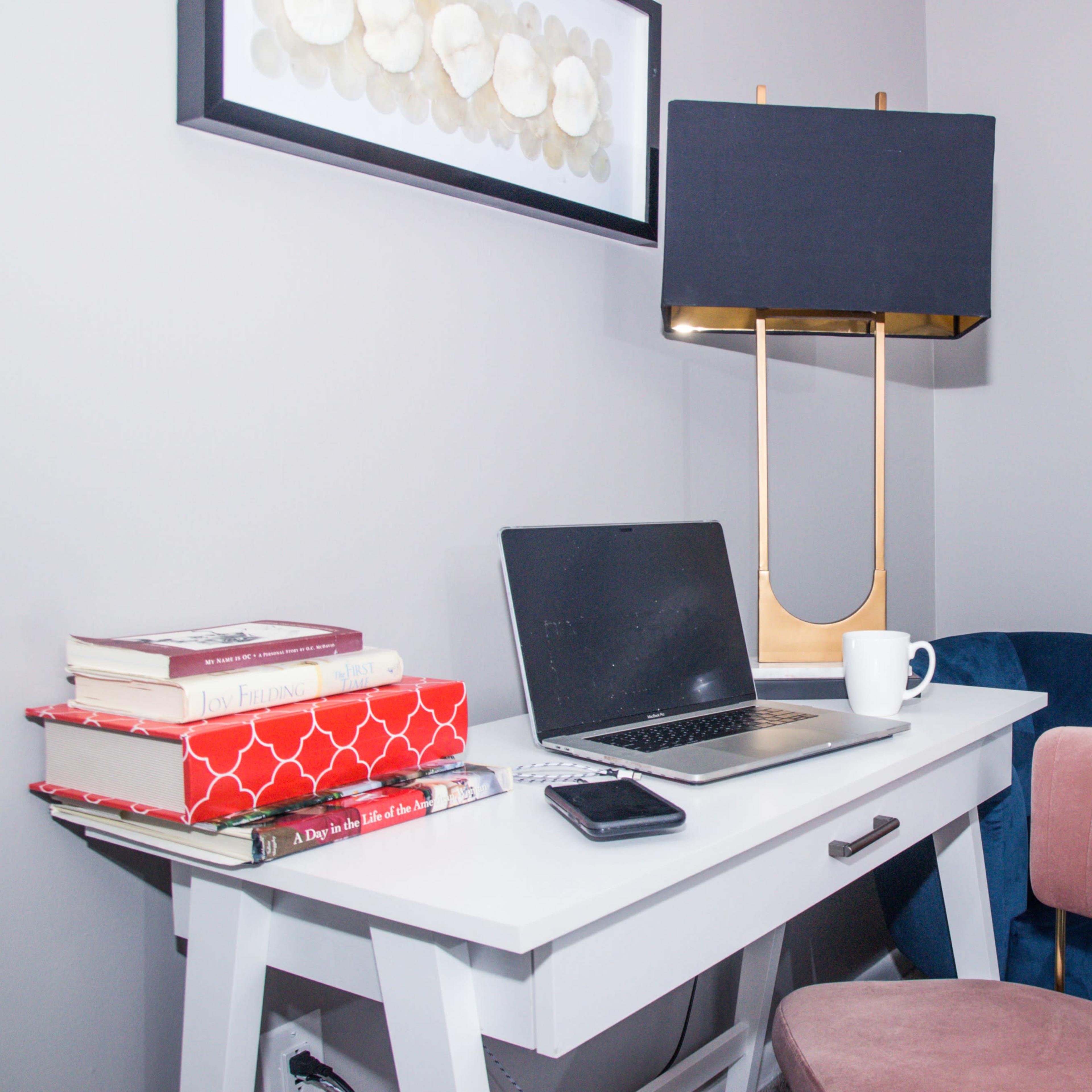 A desktop setup features a laptop, a stack of books, a coffee mug, and a table lamp, all arranged on a white desk in a small, modern workspace.