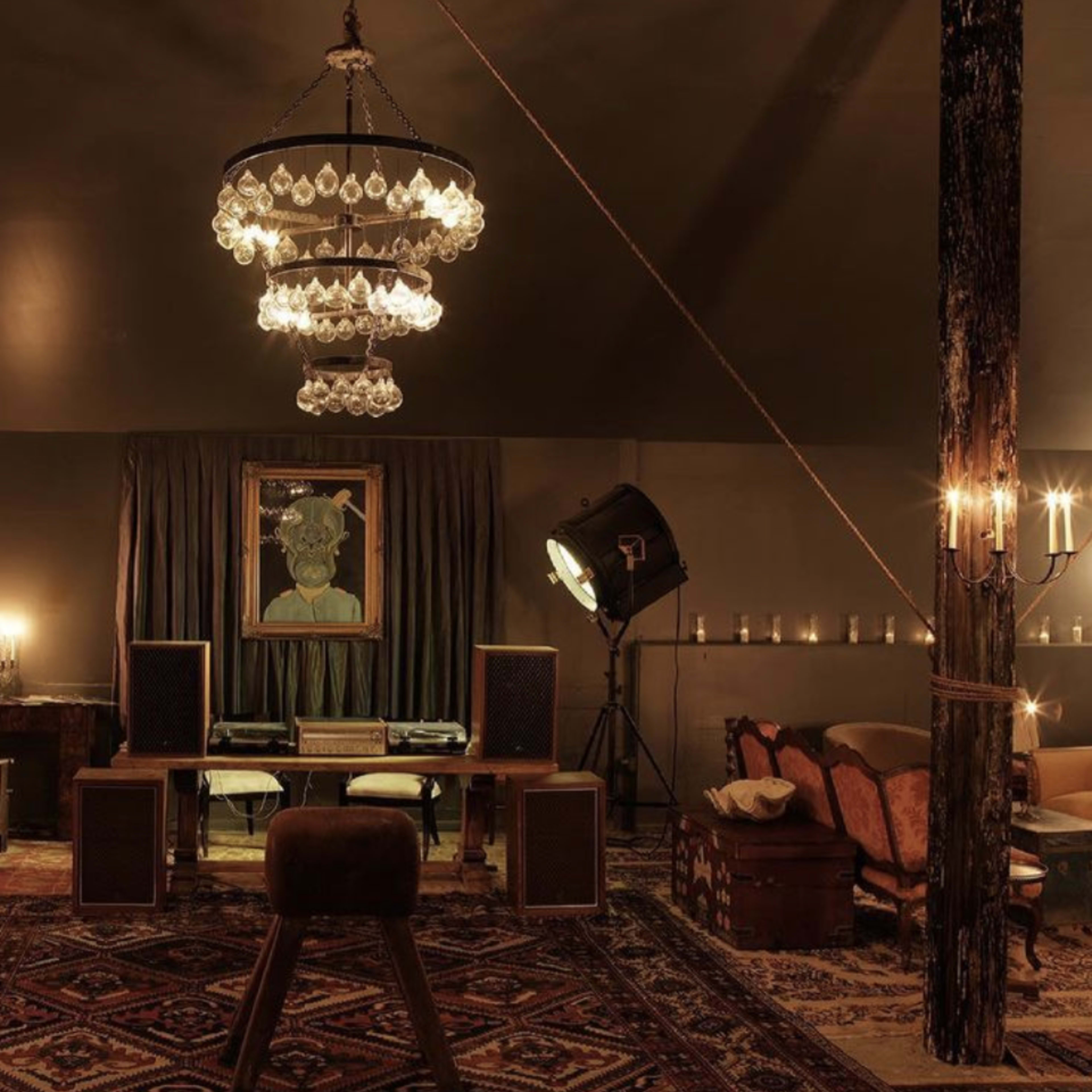 The scene features a dimly lit room with a large chandelier, wooden beams, a decorative rug, and a vintage seating area arranged around a central table with audio equipment.