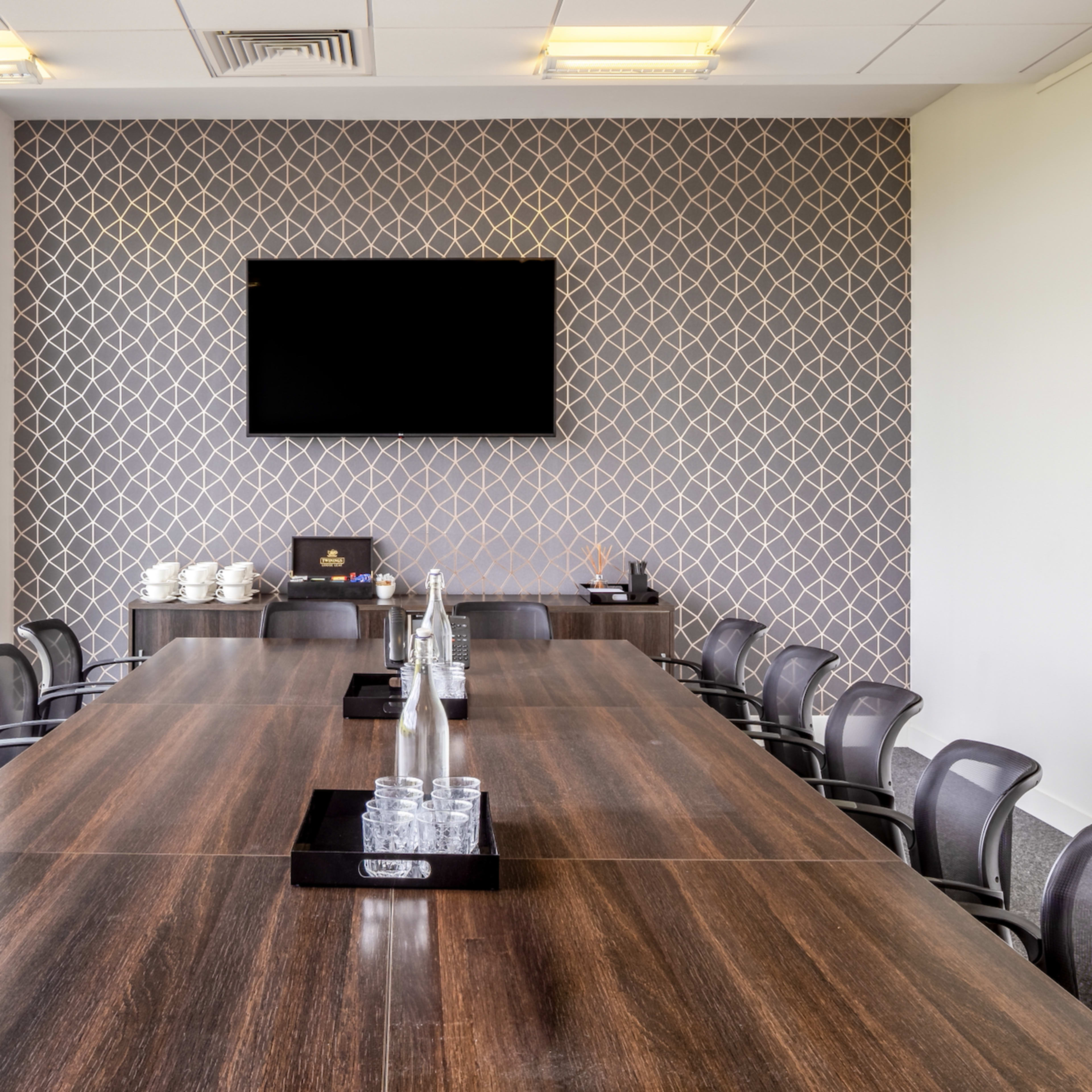 A long conference table is set in a modern meeting room with a patterned accent wall and a large screen mounted above.