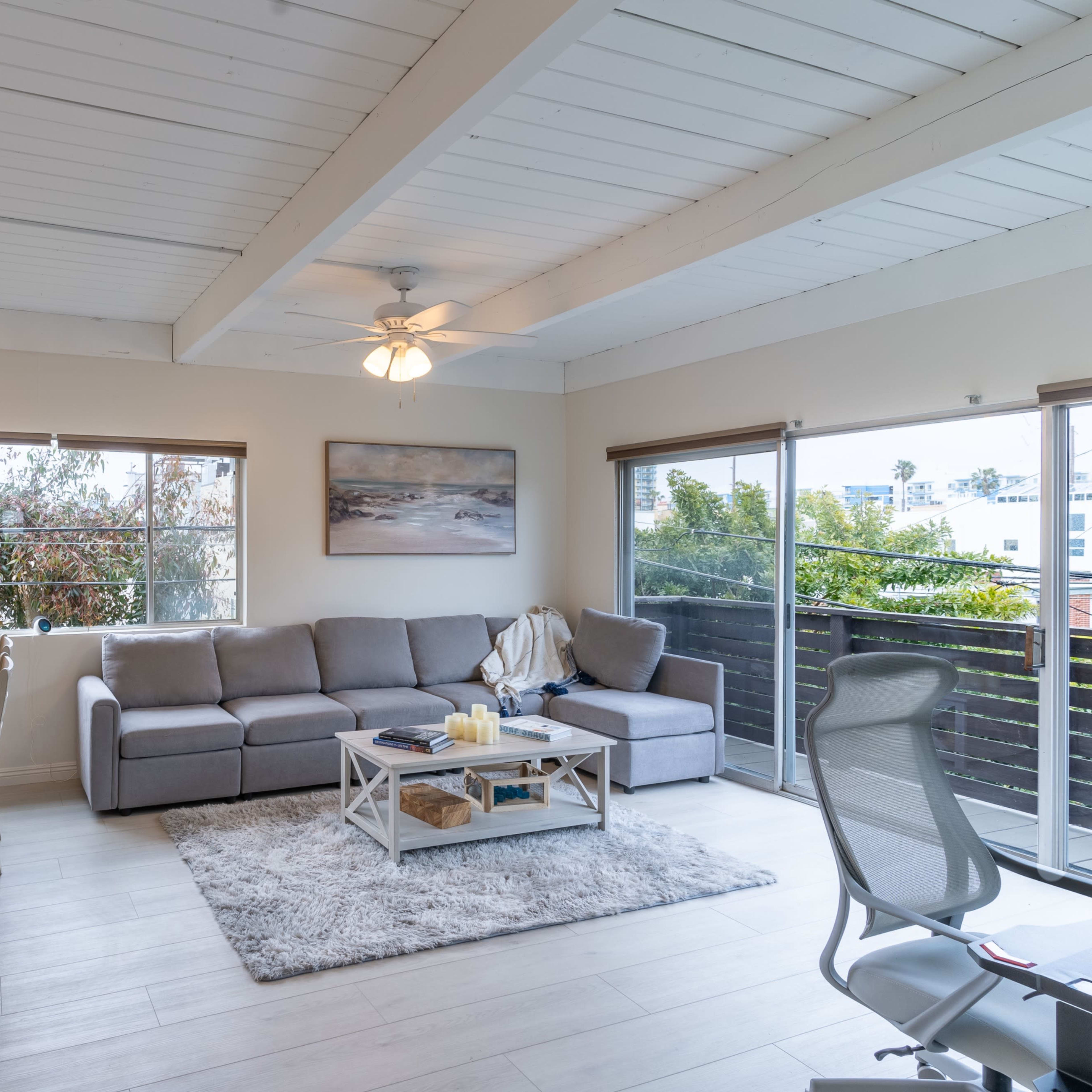 A living room and dining area with a gray sectional sofa, a light wooden table, and large windows providing natural light.