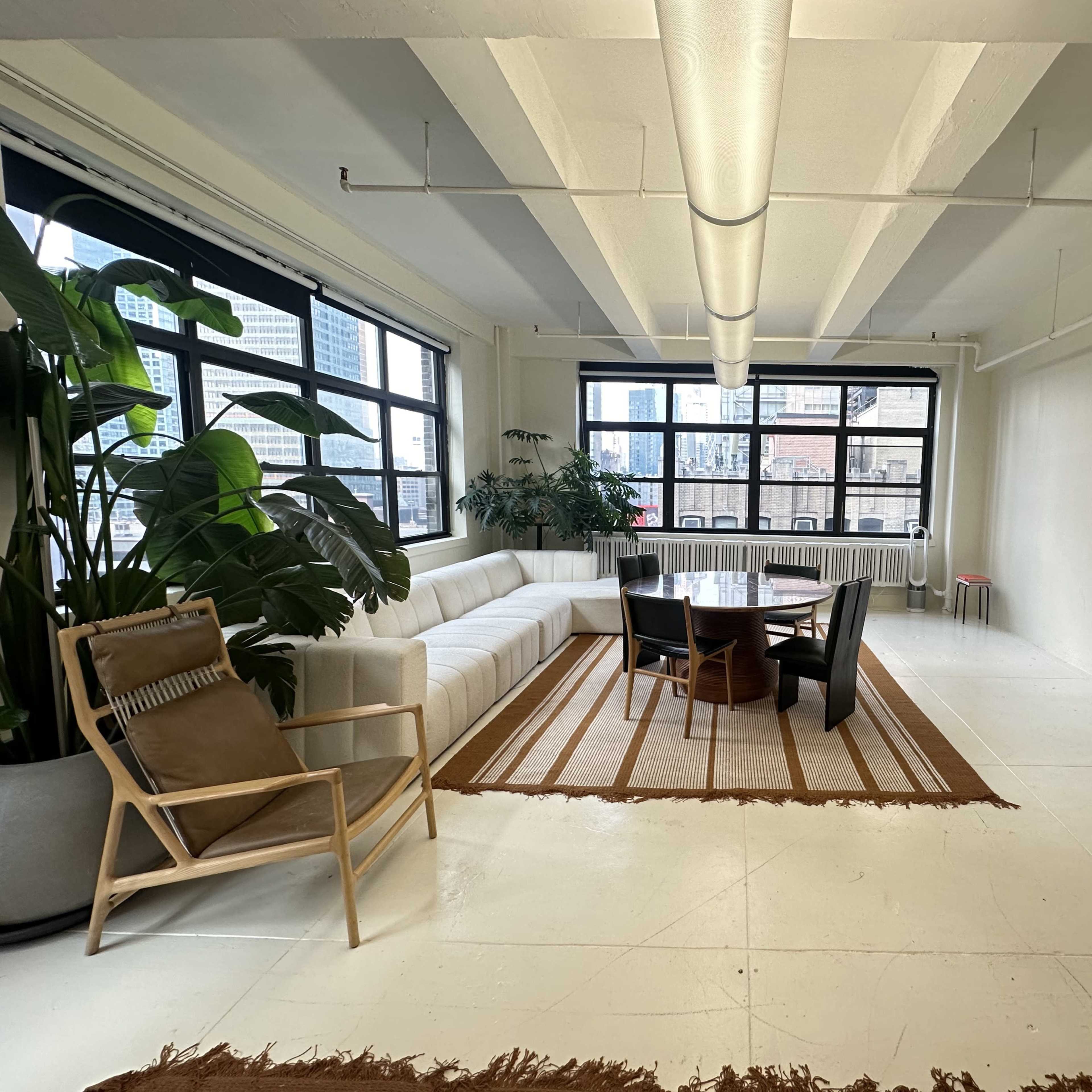 A spacious, well-lit room features a large sectional sofa, a round dining table with chairs, and potted plants near large windows overlooking a cityscape.