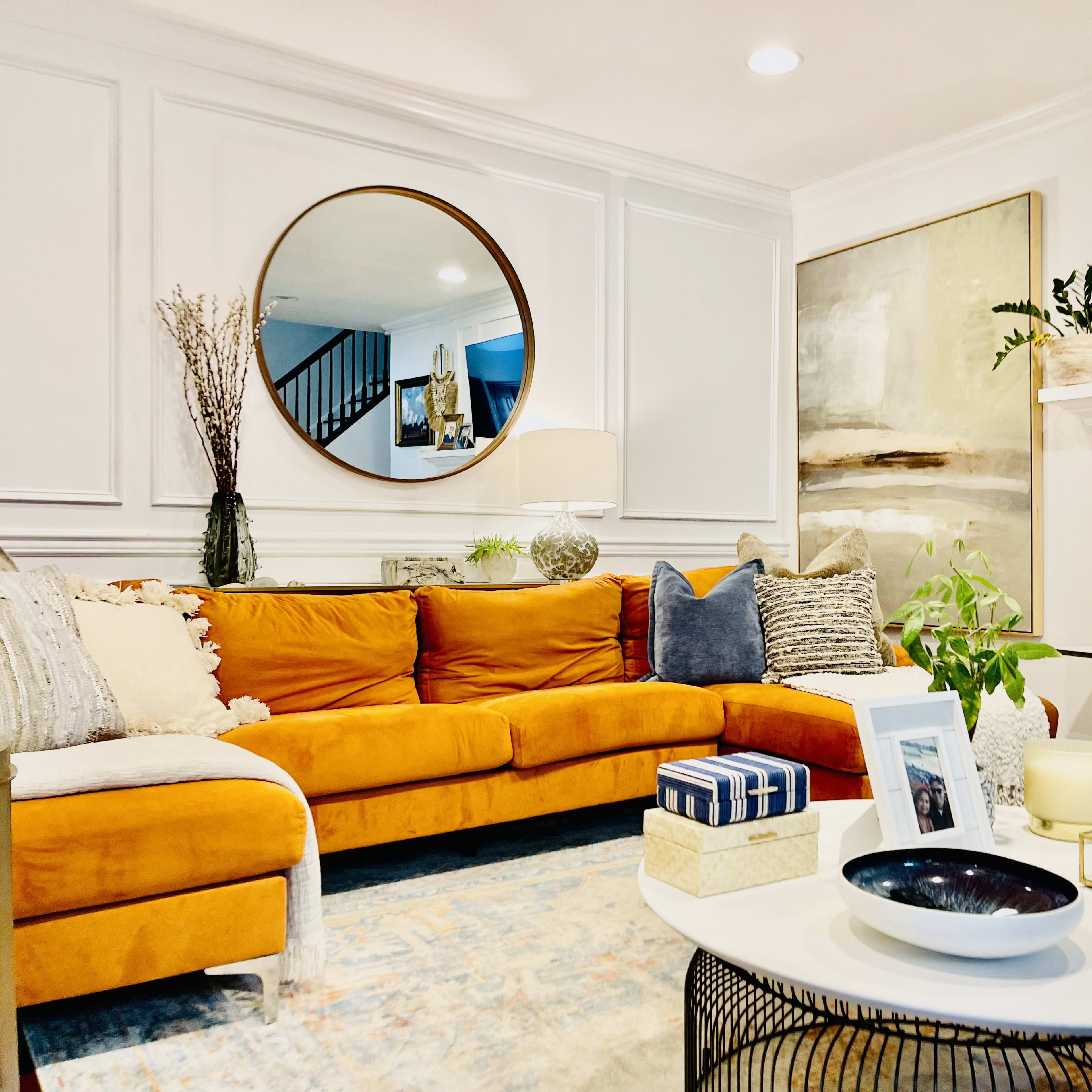 A modern living room features a large orange sectional sofa, a round mirror above a decorative fireplace, and various art pieces and plants arranged thoughtfully throughout the space.